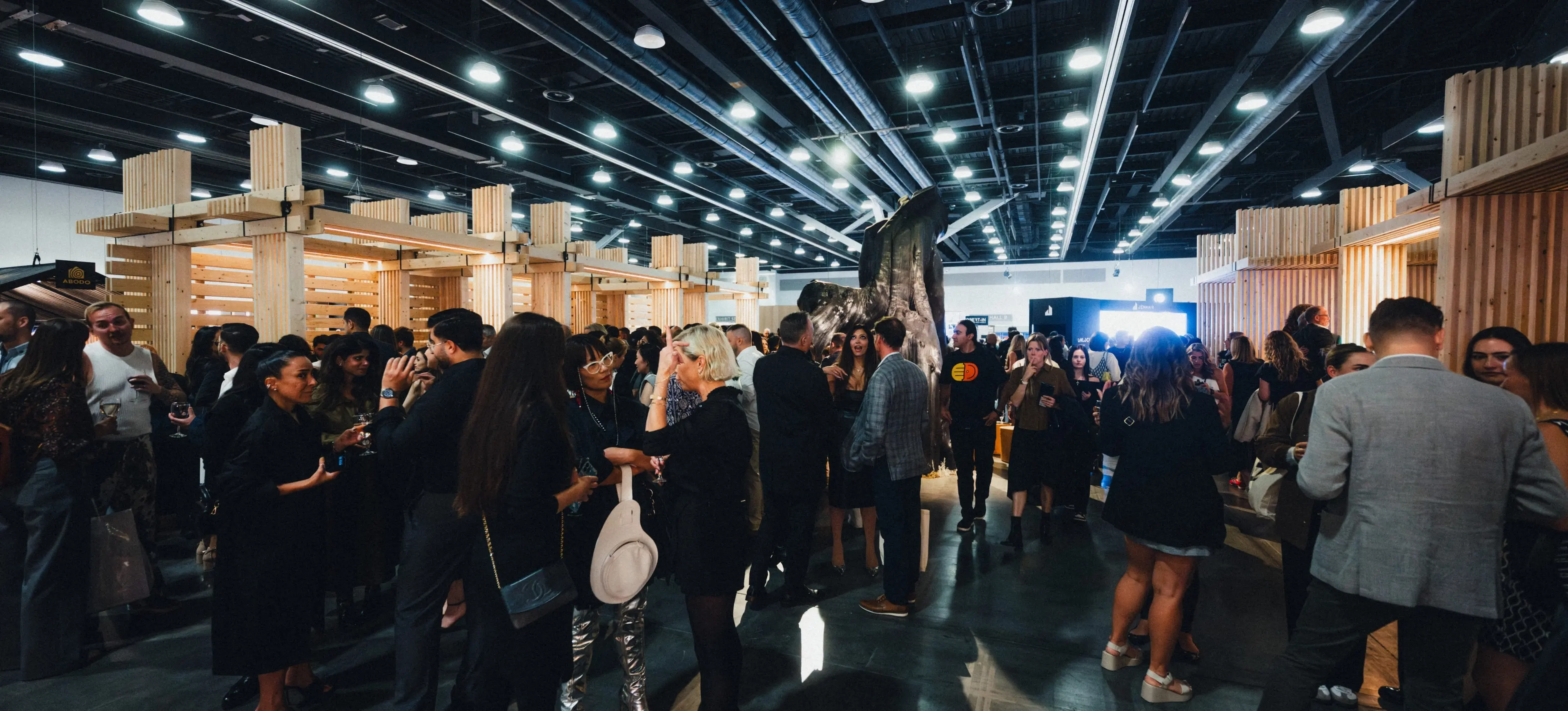 Crowd of people socializing in a large indoor event space with wooden structures and bright ceiling lights.