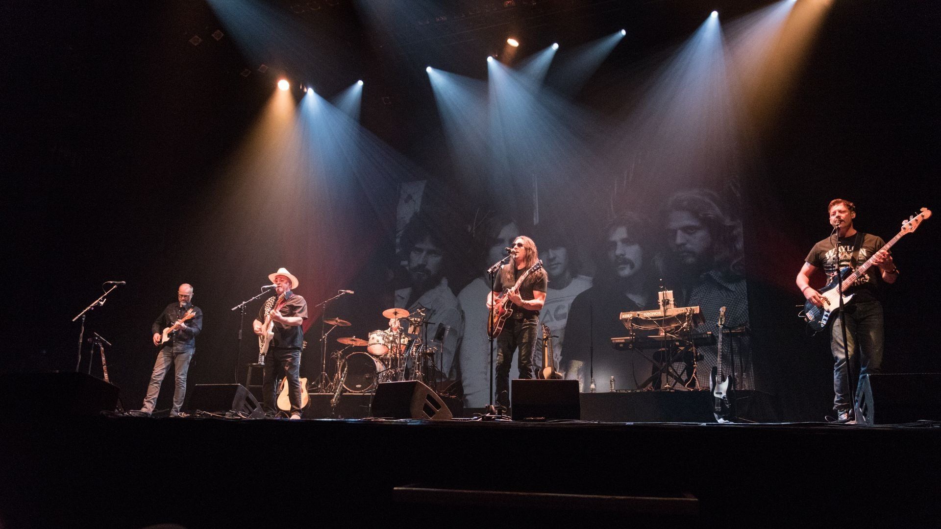 Five musicians performing on stage with guitars, drums, and keyboard under spotlights and a black-and-white backdrop.