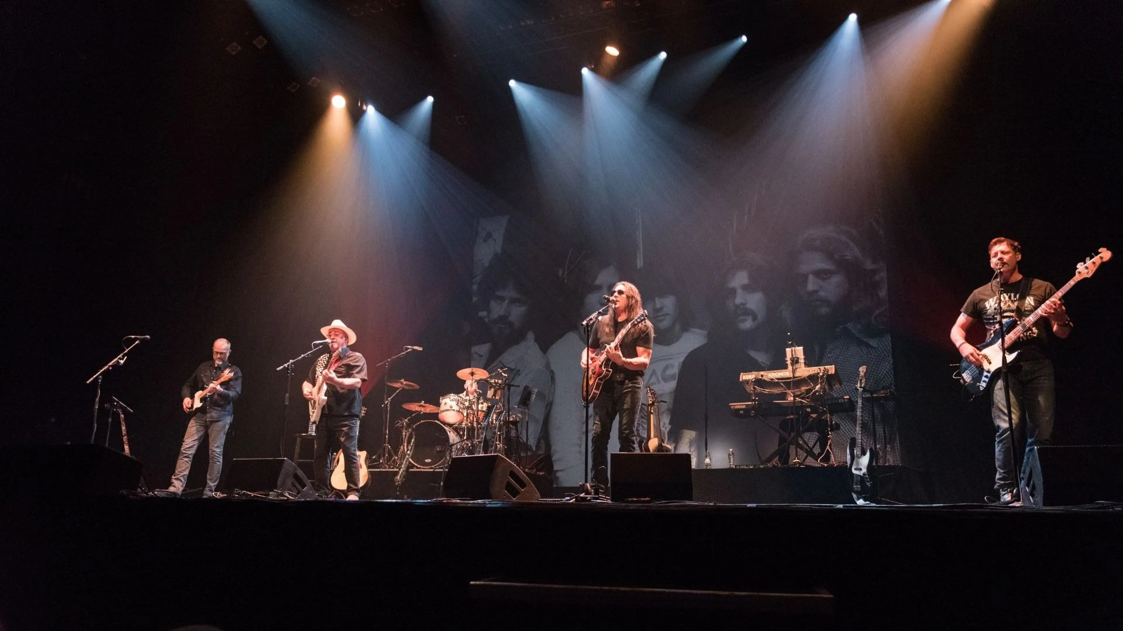 Five musicians performing on stage with guitars, drums, and keyboard under spotlights and a black-and-white backdrop.