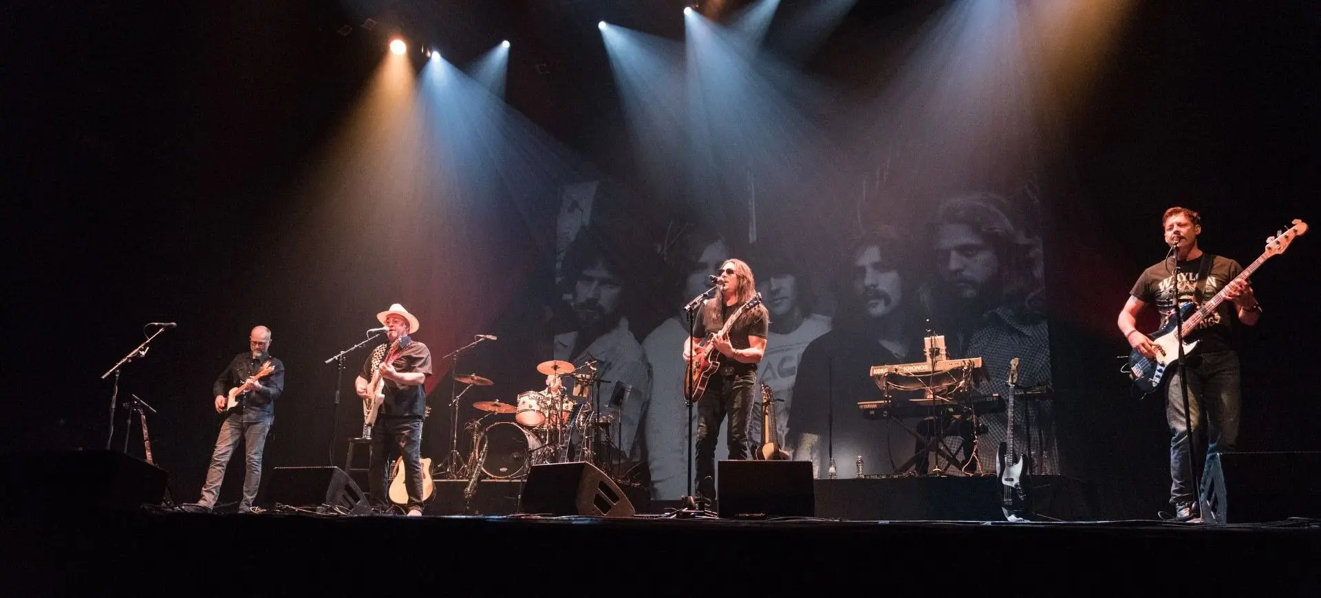 Five musicians performing on stage with guitars, drums, and keyboard under spotlights and a black-and-white backdrop.