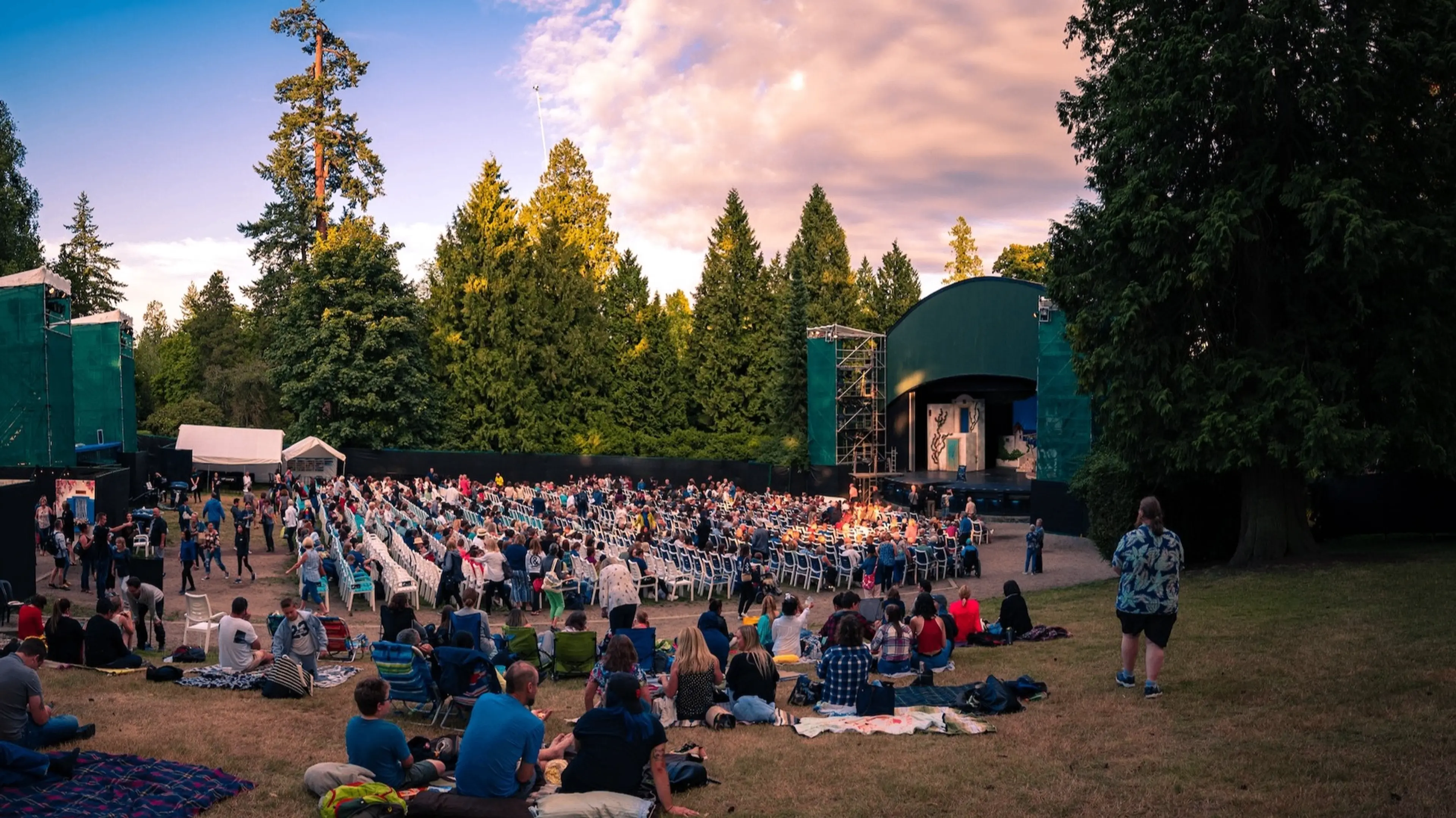 Outdoor amphitheater with a crowd seated on chairs and blankets watching a stage surrounded by tall trees at sunset.