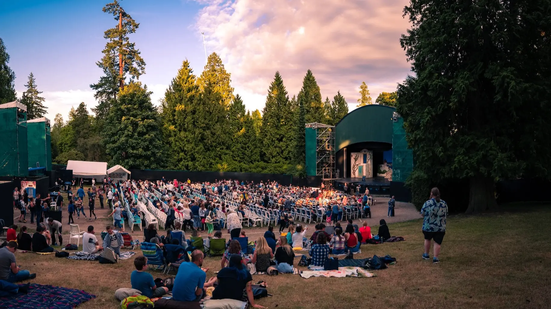 Outdoor amphitheater with a crowd seated on chairs and blankets watching a stage surrounded by tall trees at sunset.