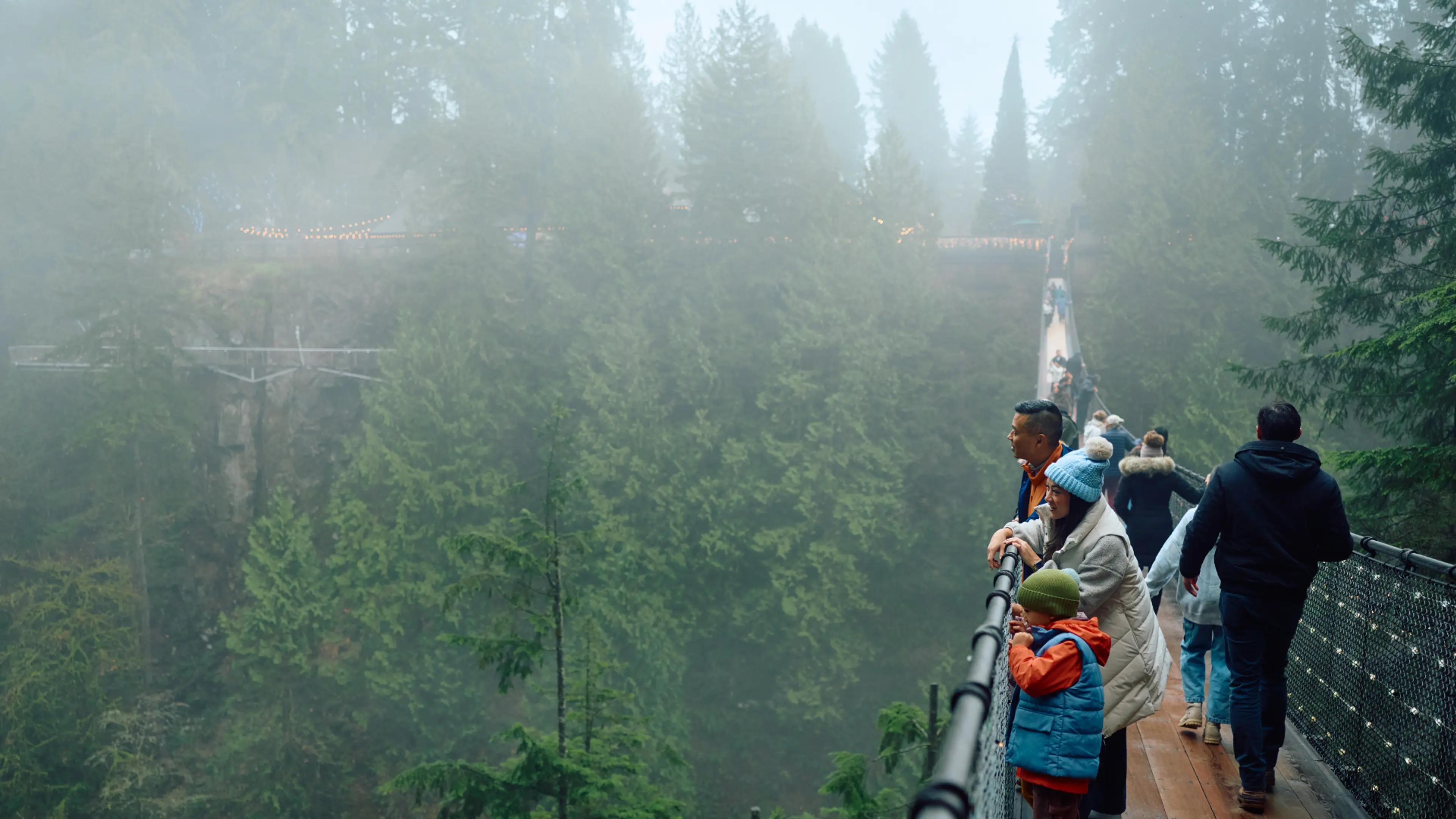 People standing and walking on a foggy suspension bridge surrounded by tall evergreen trees.