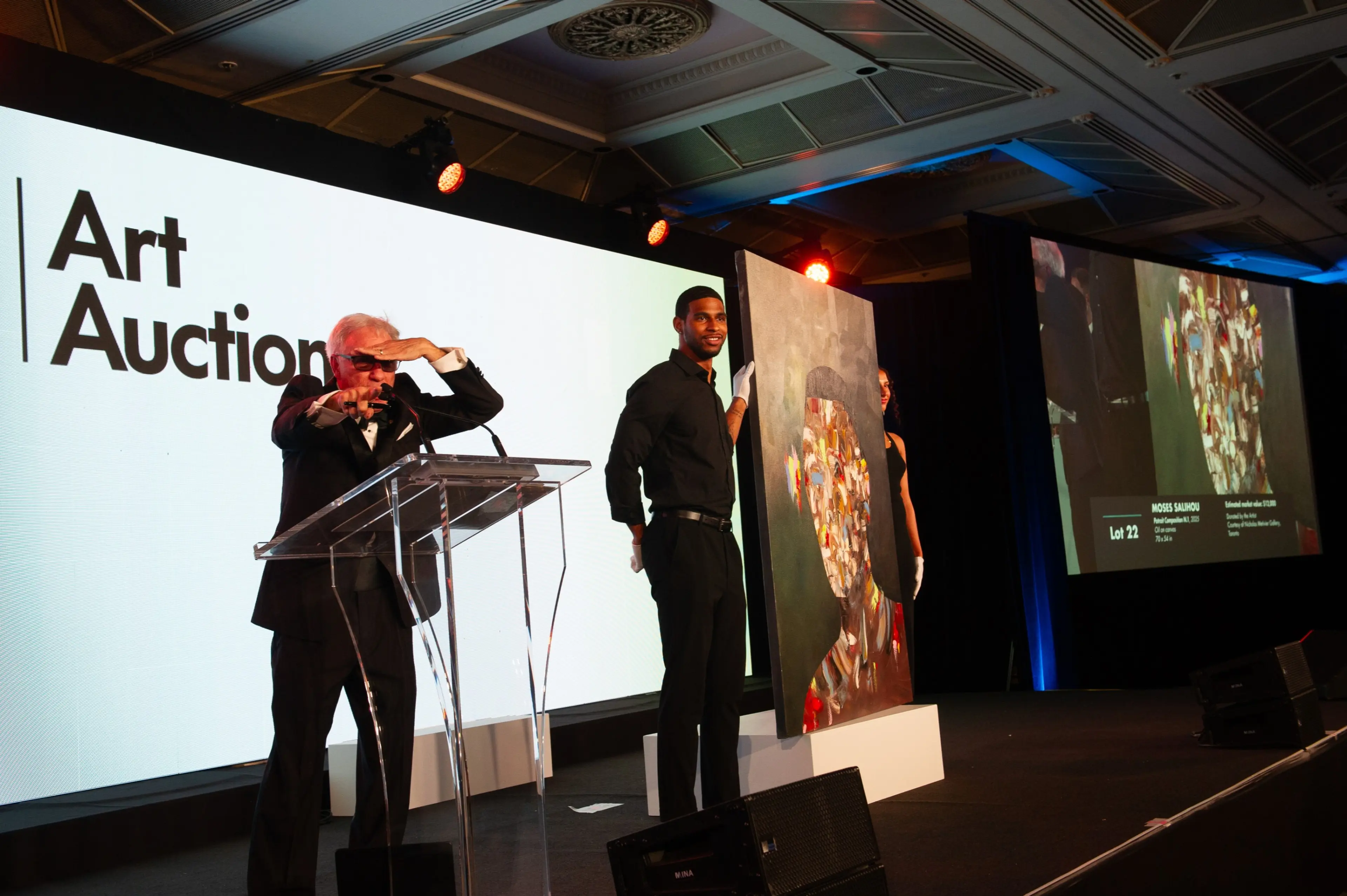 An auctioneer at a podium gestures while two people hold a large colorful portrait painting on stage at an art auction.