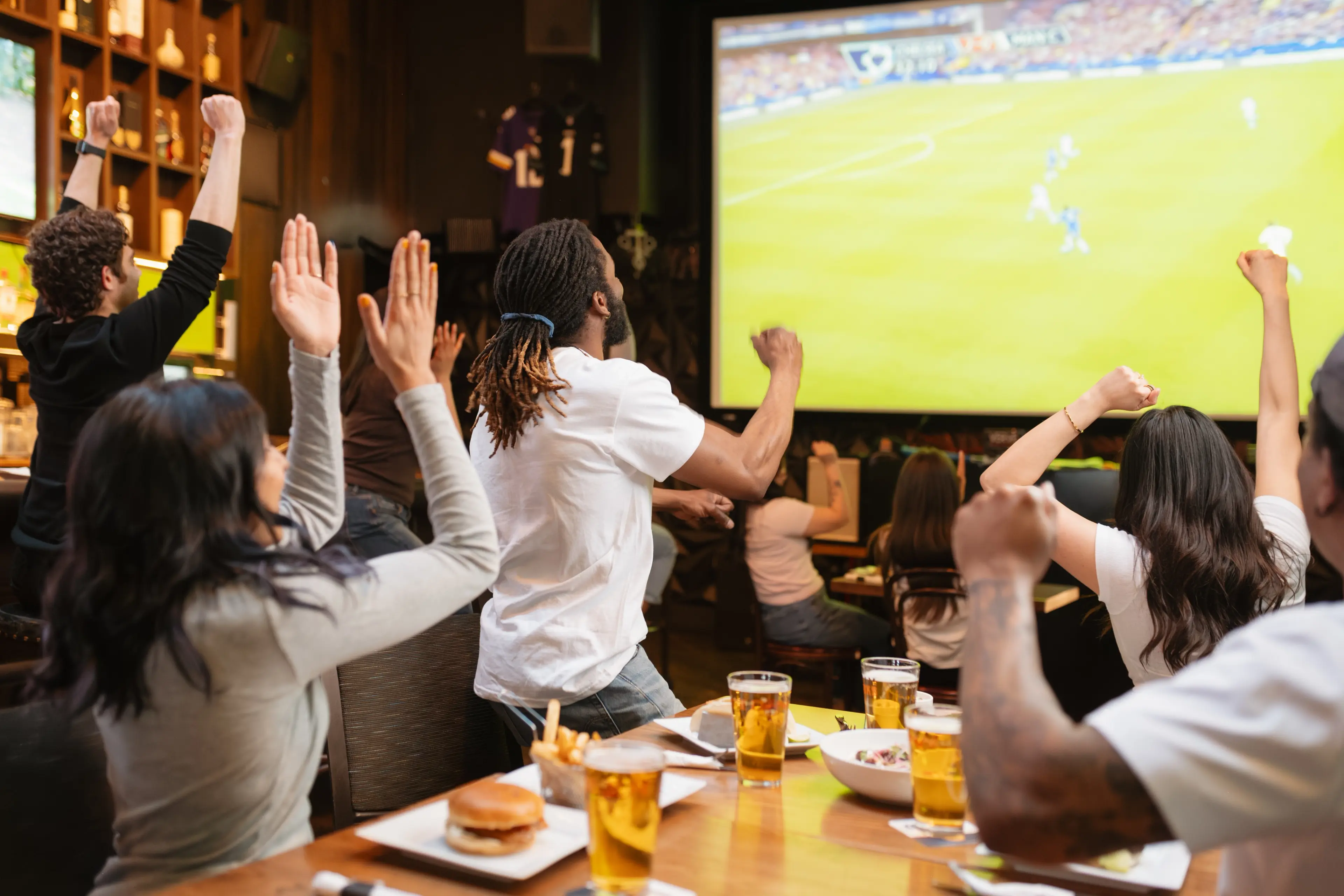Group of people in a bar cheering and raising hands while watching a soccer game on a large screen.