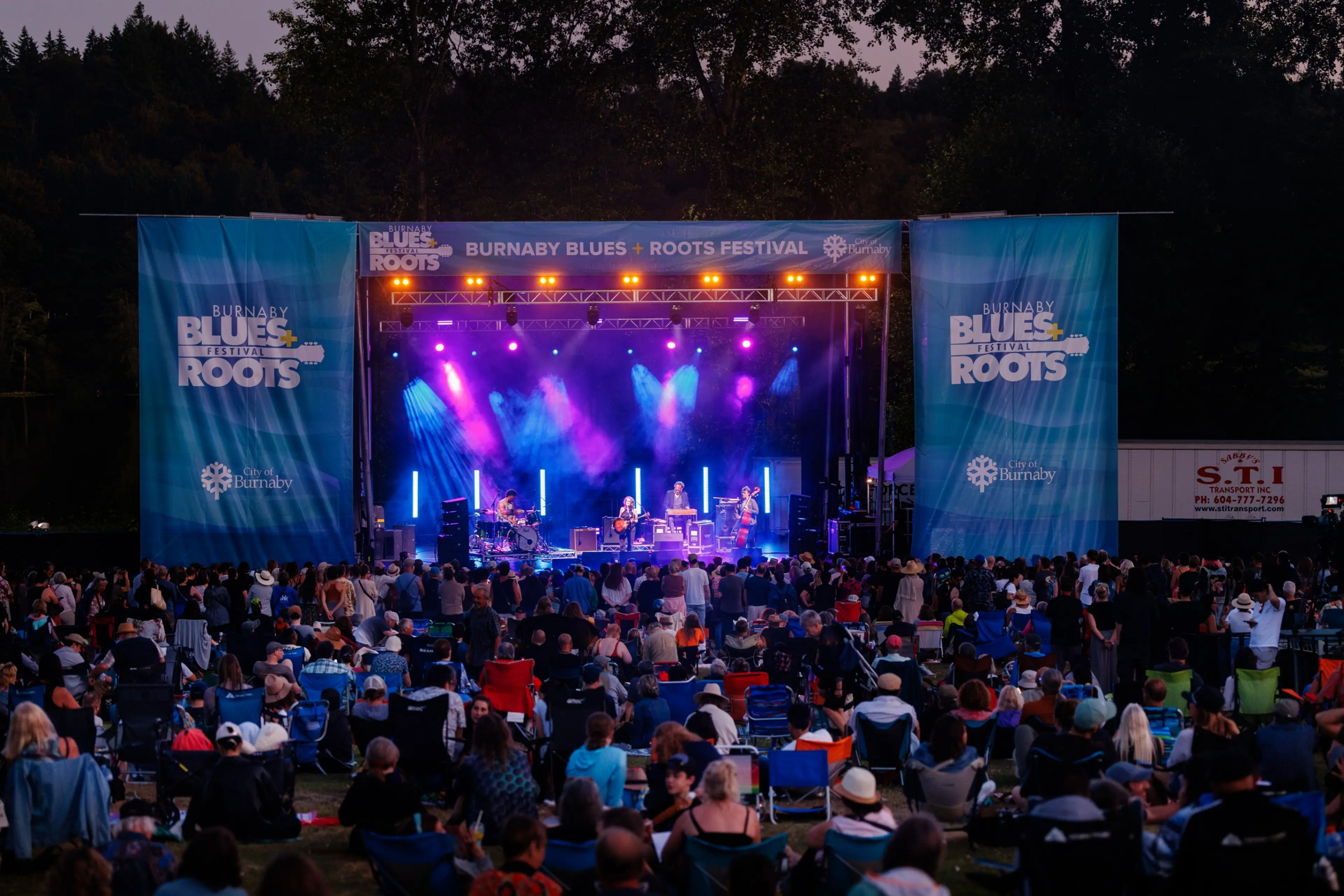 Outdoor concert stage at Burnaby Blues + Roots Festival with band performing and large seated audience at dusk.