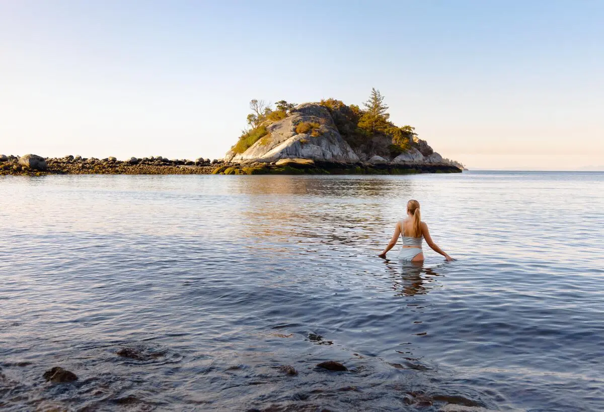 A person in a swimsuit wading into calm water with a rocky island and clear sky in the background.