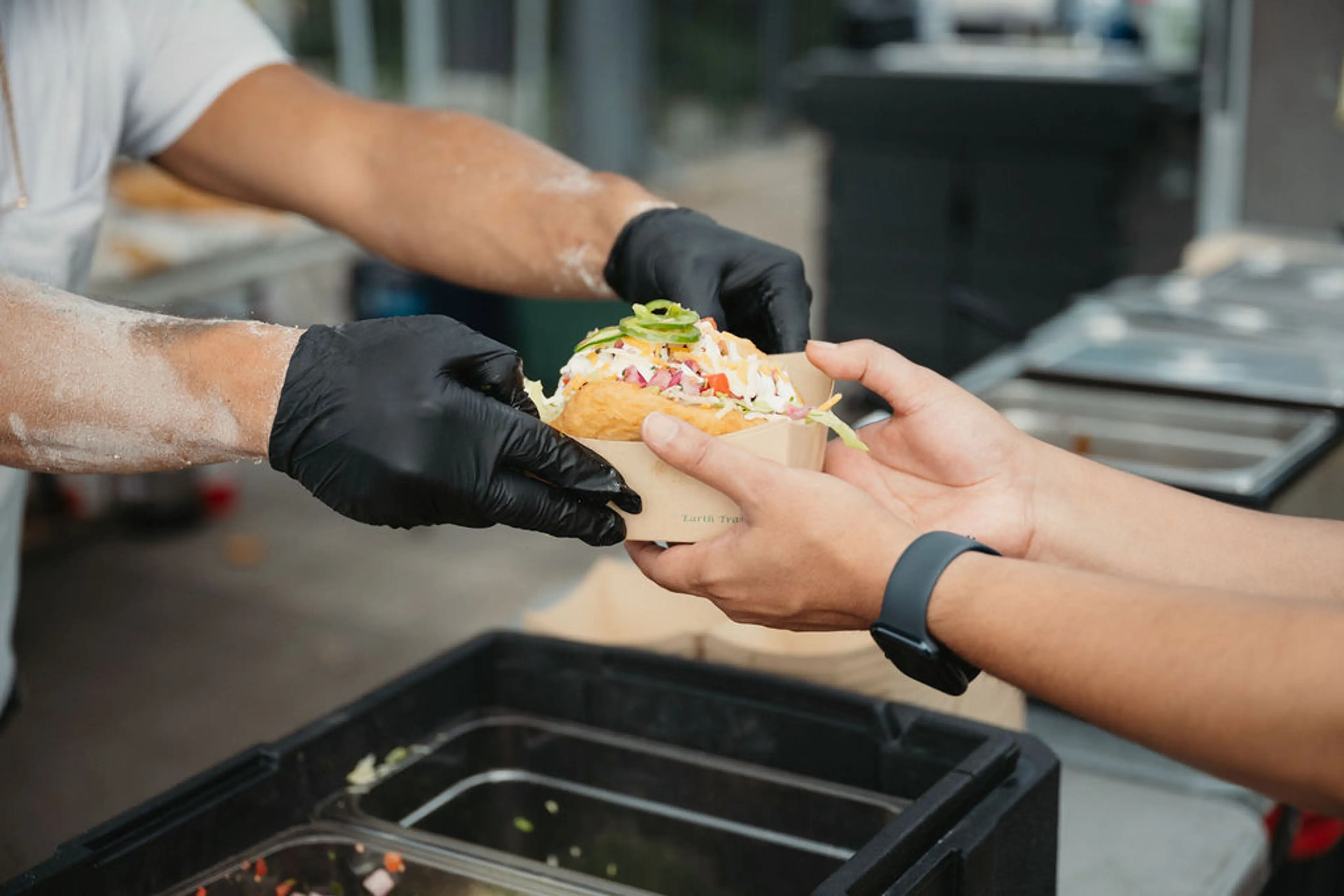 Person wearing black gloves handing a sandwich with toppings to another person wearing a black wristband.