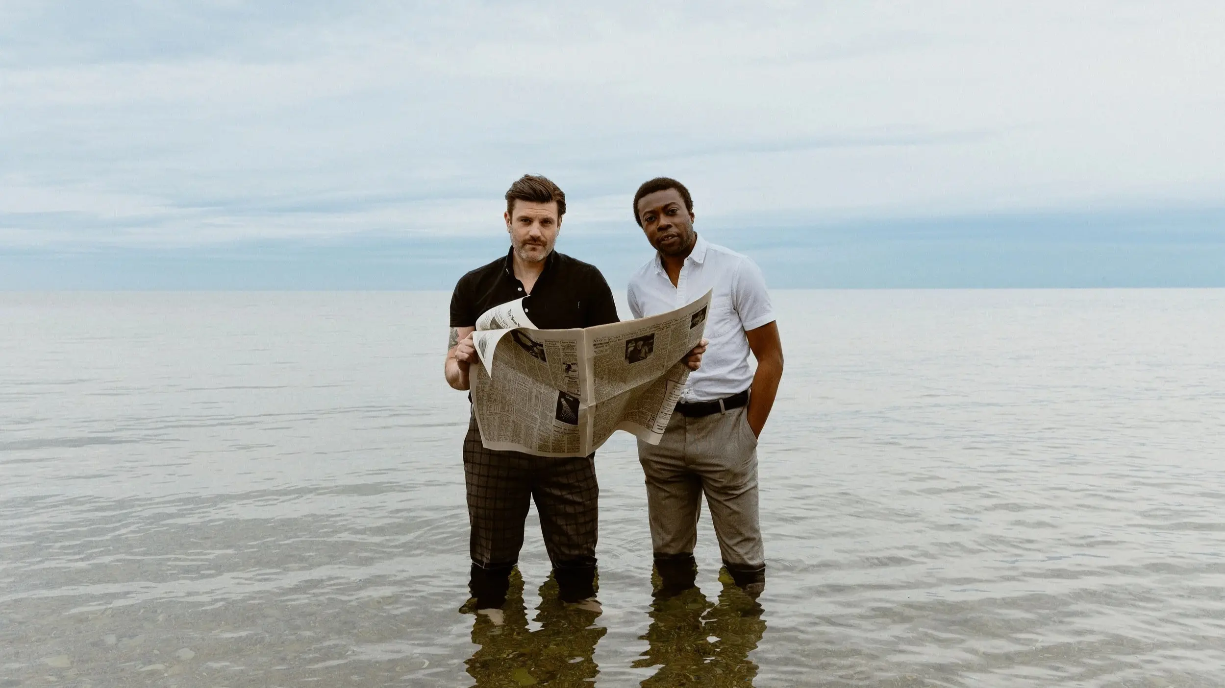 Two people standing in shallow water at the beach, one holding a newspaper, both looking at the camera.