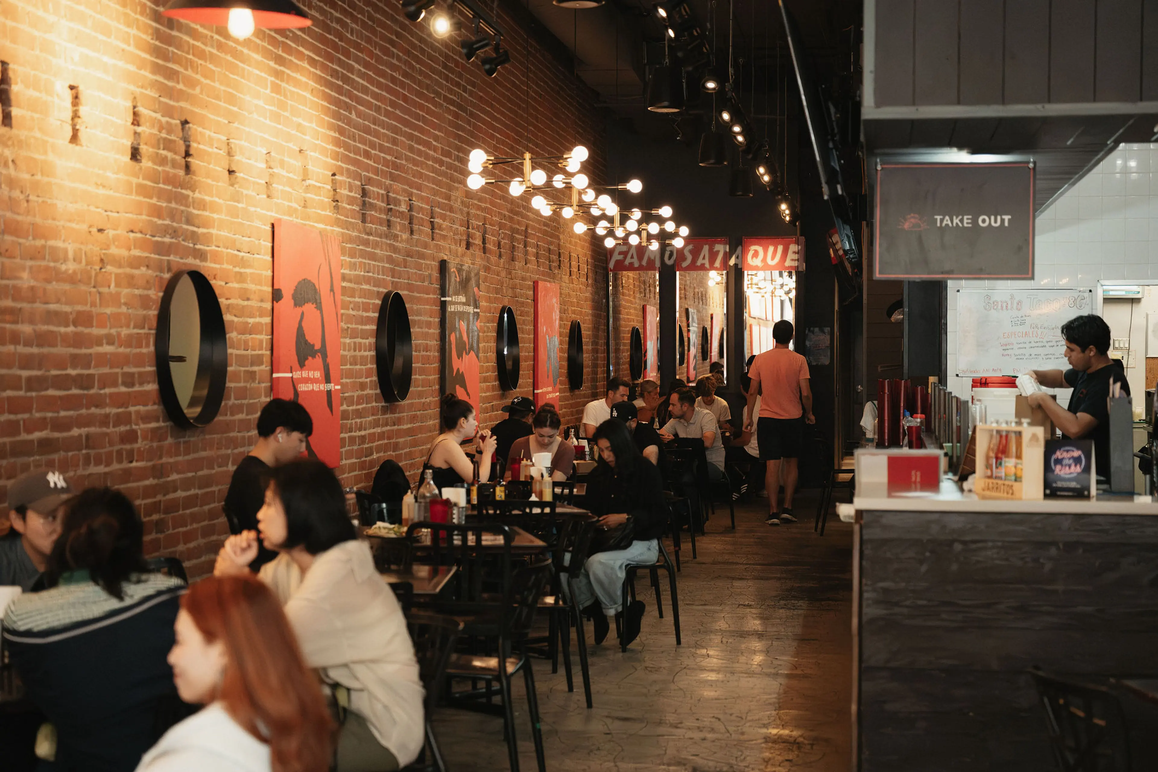 People sitting and eating at tables along a brick wall with round mirrors and red posters in a restaurant with a takeout counter on the right.