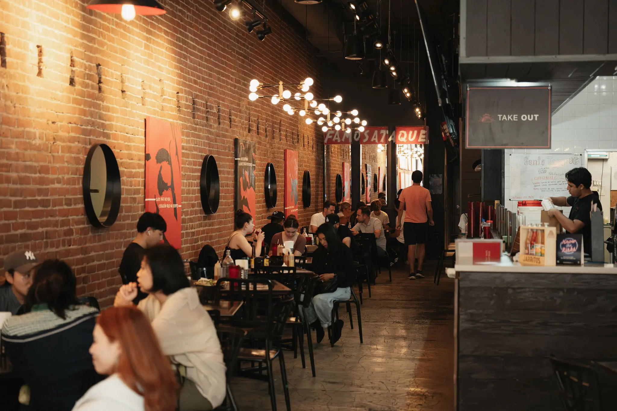 People sitting and eating at tables along a brick wall with round mirrors and red posters in a restaurant with a takeout counter on the right.