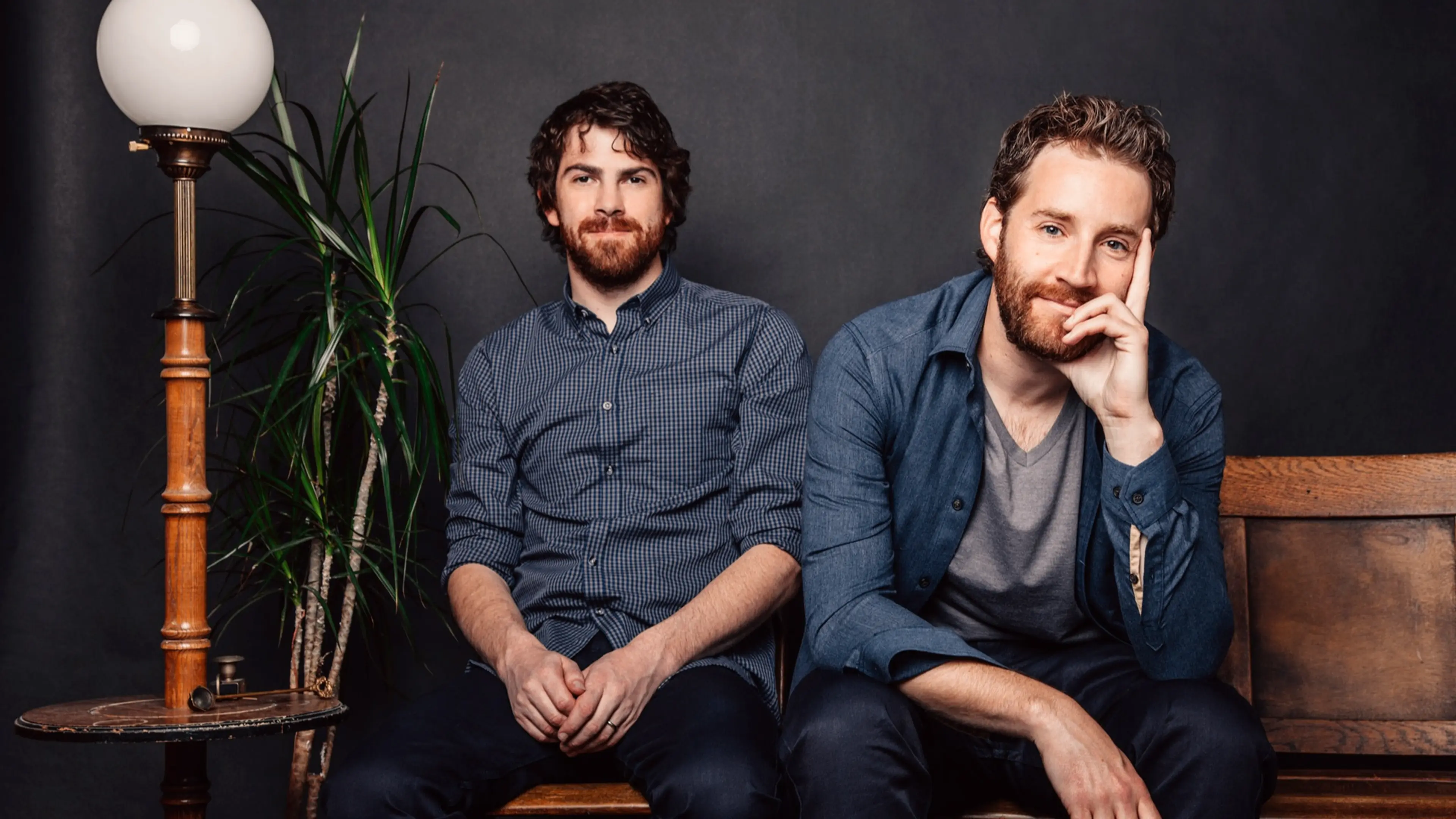 Two seated people with beards and casual shirts against a dark background, next to a wooden lamp and a tall plant.