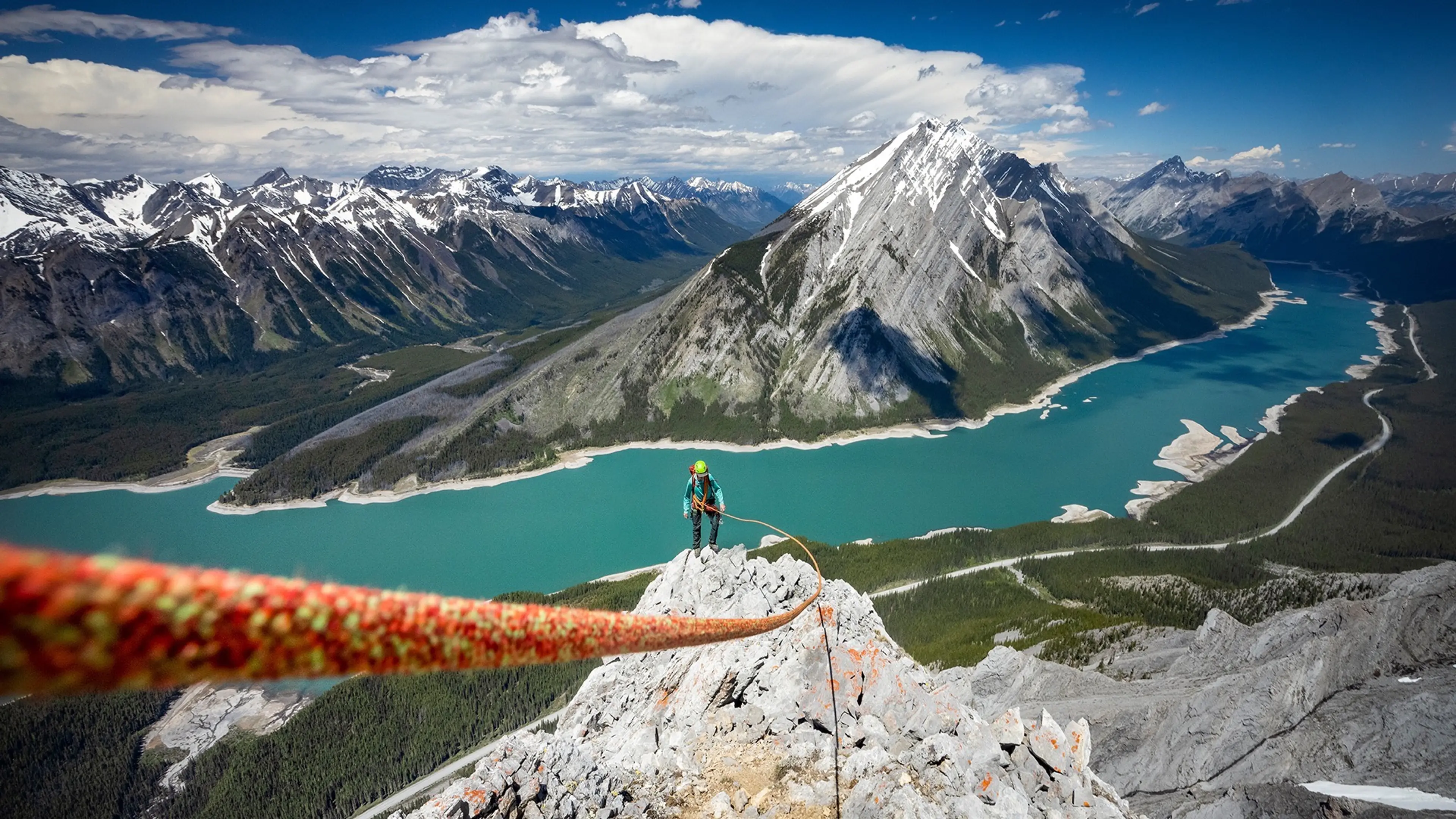 A person climbing a rocky mountain peak with a safety rope, overlooking a turquoise river and snow-capped mountains under a partly cloudy sky.