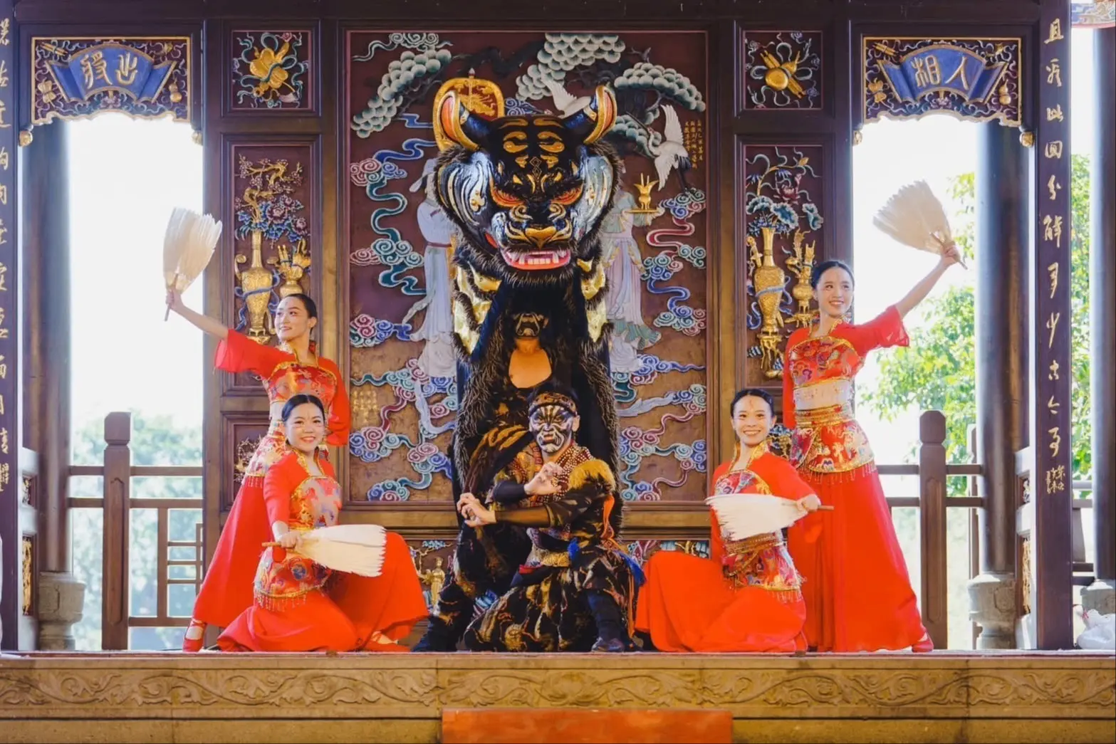 Four people in red traditional attire holding fans pose around two performers in a black and orange tiger costume on a decorated stage.