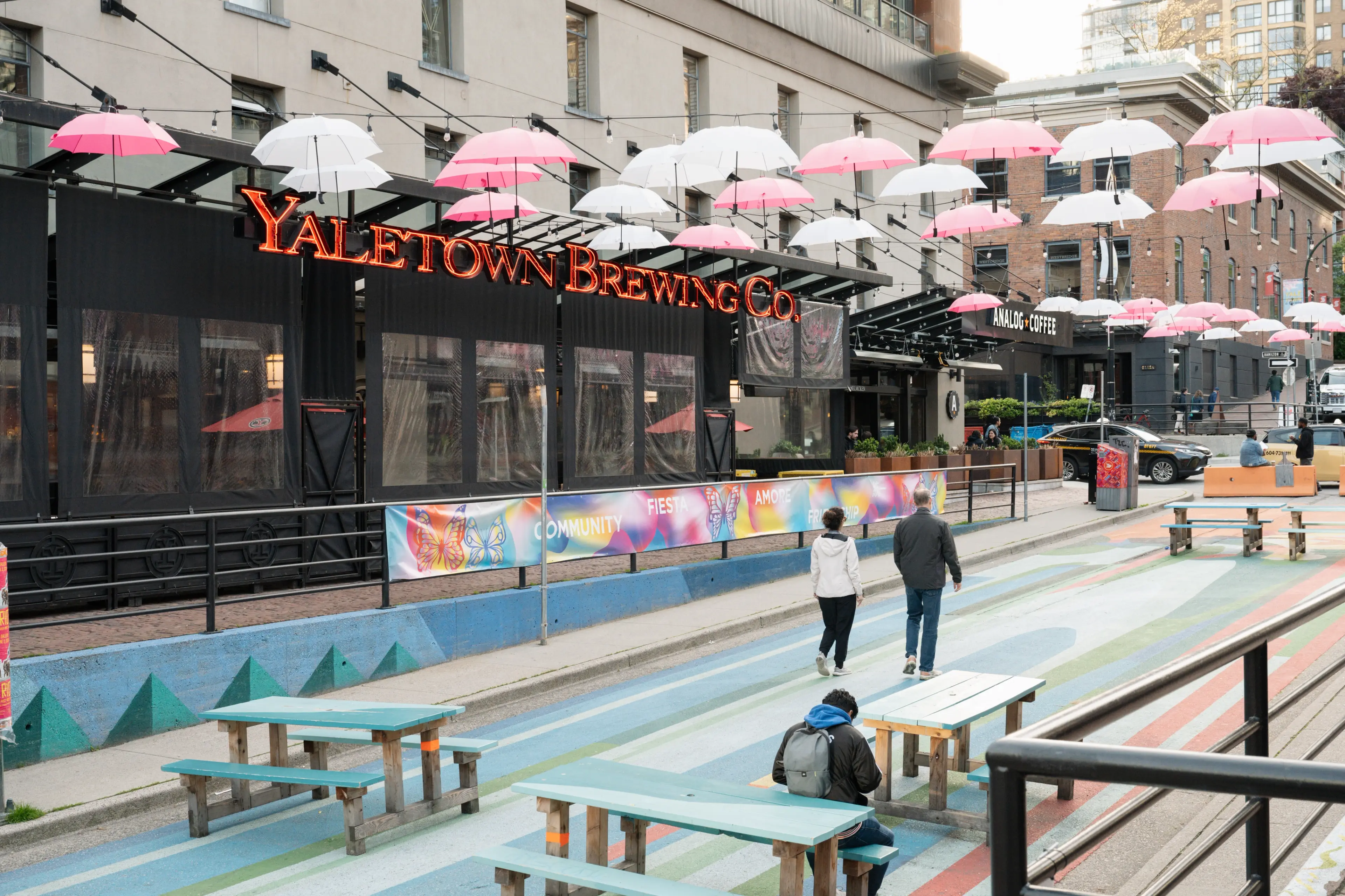 Outdoor seating area with colorful striped ground, wooden benches, and pink and white umbrellas hanging above near Yaletown Brewing Co.
