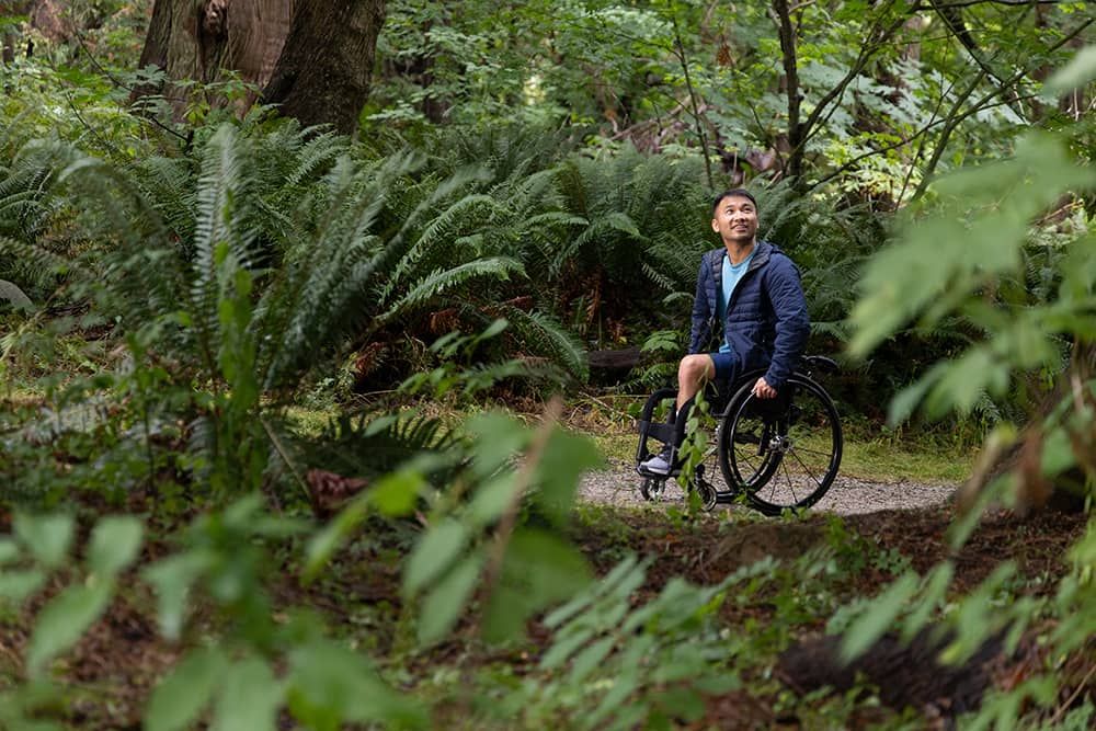 Person in a wheelchair on a forest trail surrounded by green ferns and trees, looking up and smiling.