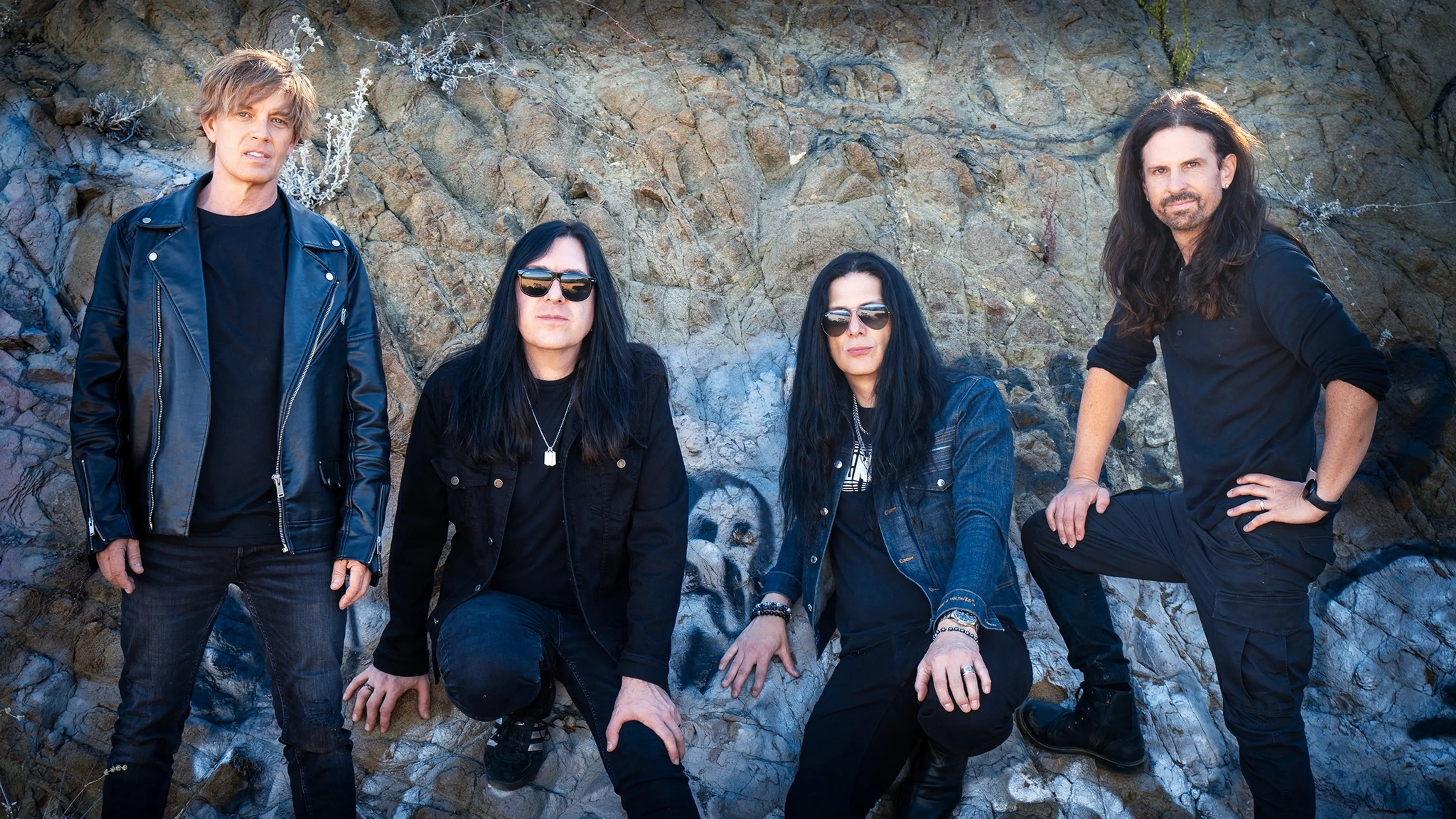 Four people with long and short hair wearing dark clothing and sunglasses posing against a rocky wall with graffiti.