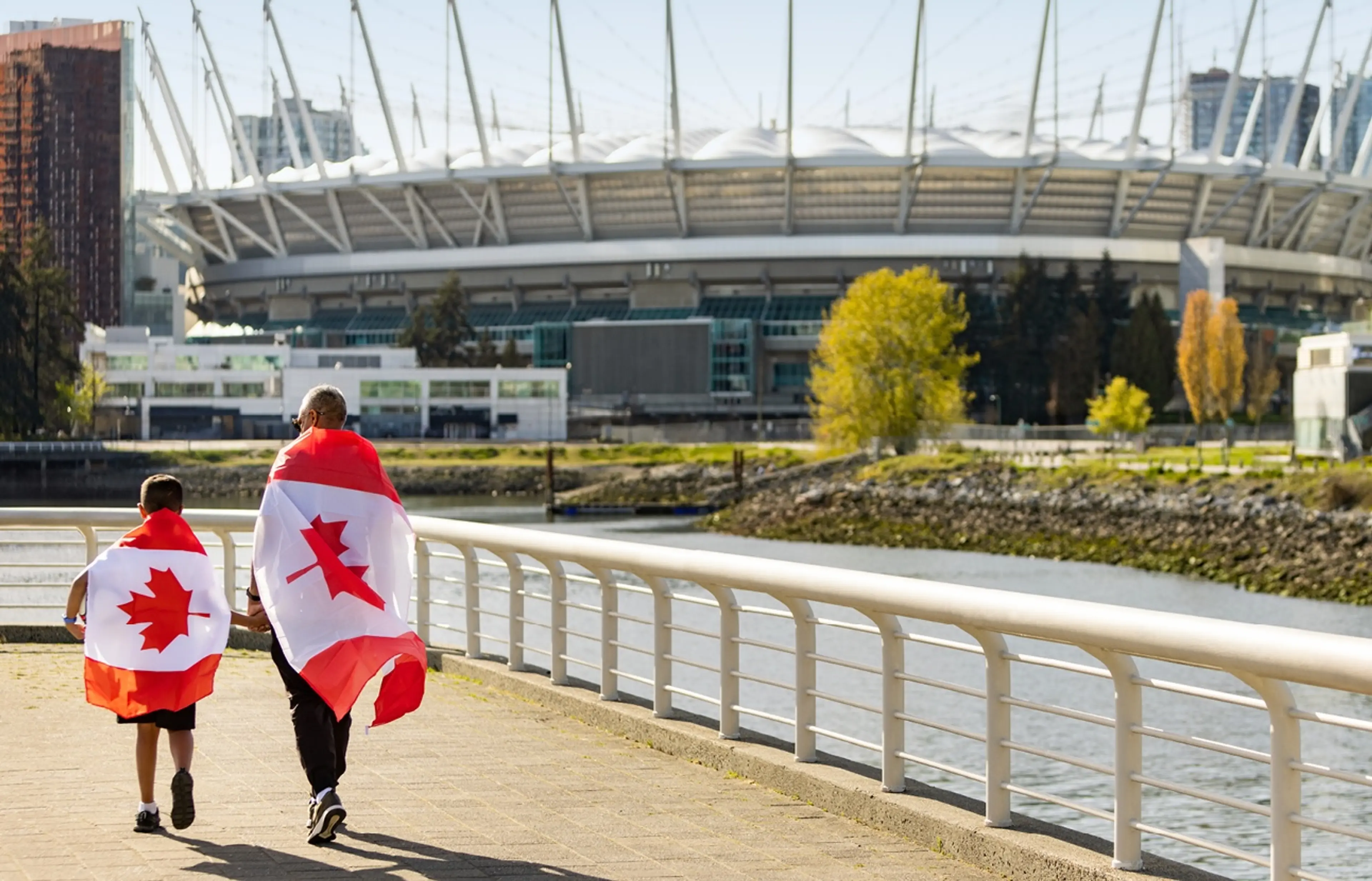 Two people walking along a waterfront path, each draped in a Canadian flag, with a stadium and trees in the background.
