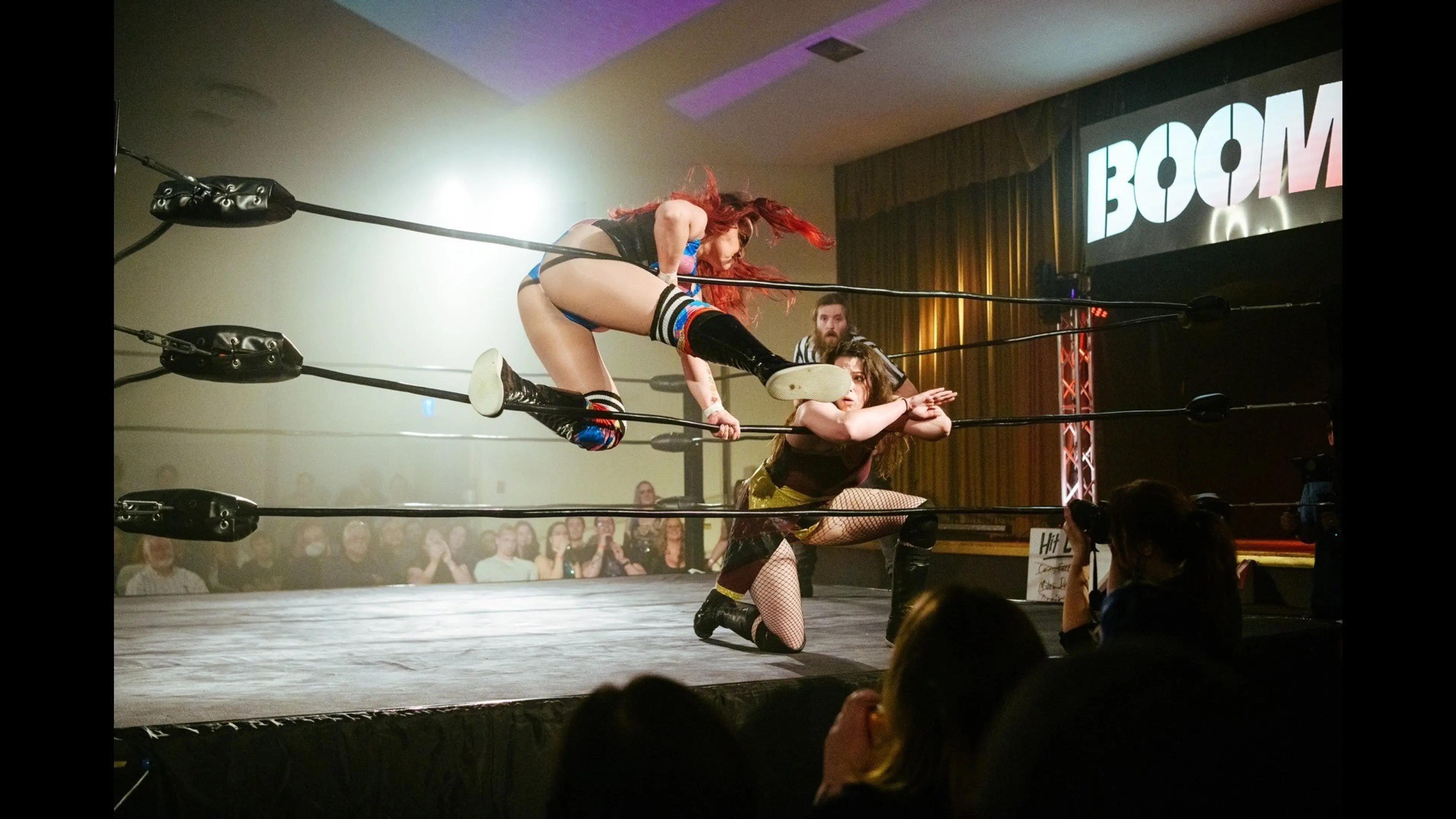 A wrestler in black and blue jumps over the ropes toward a crouching opponent in a wrestling ring with a referee and audience in the background.
