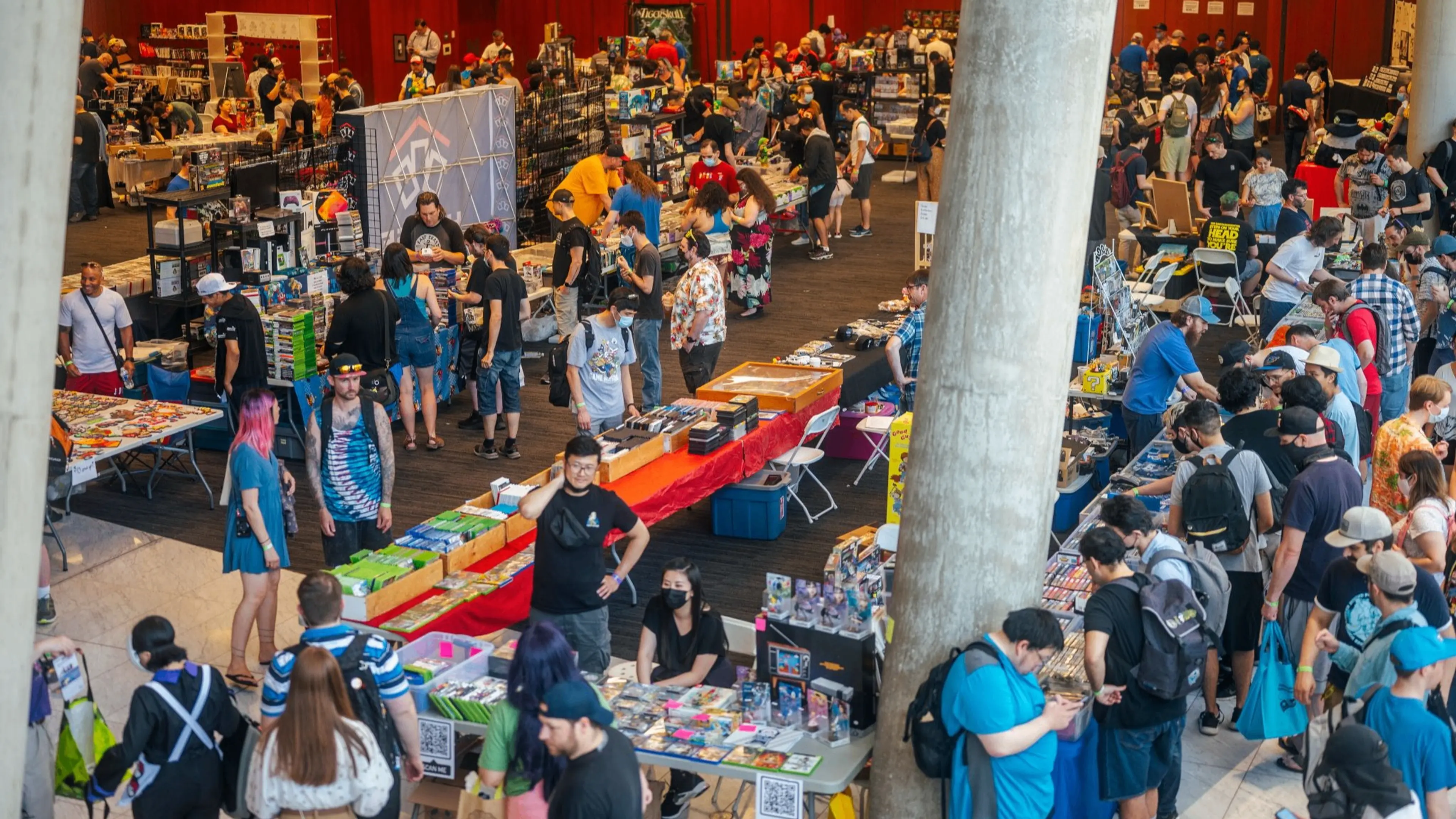 Crowded indoor market or convention with many people browsing tables filled with various items and merchandise.