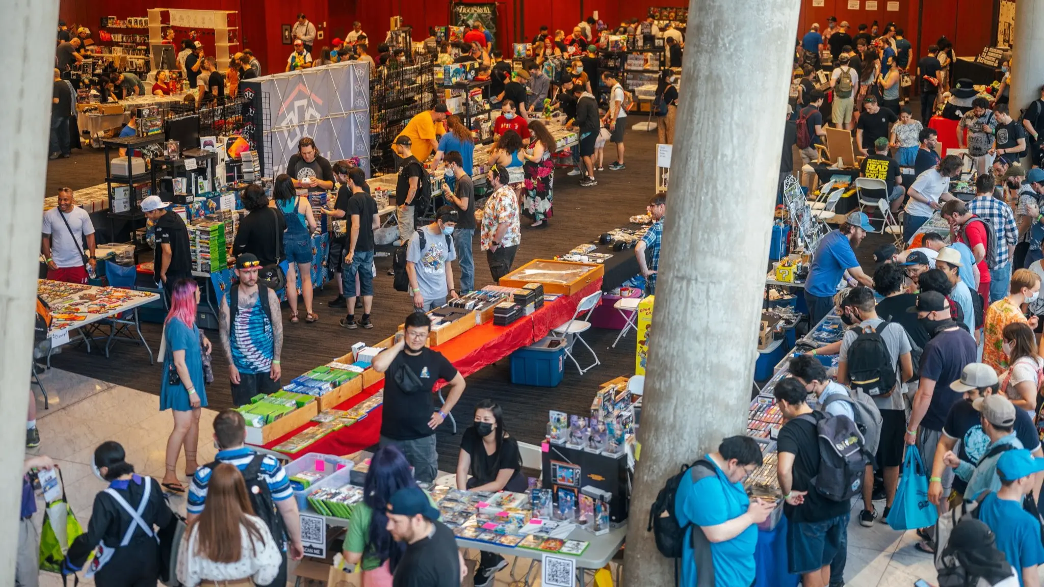Crowded indoor market or convention with many people browsing tables filled with various items and merchandise.