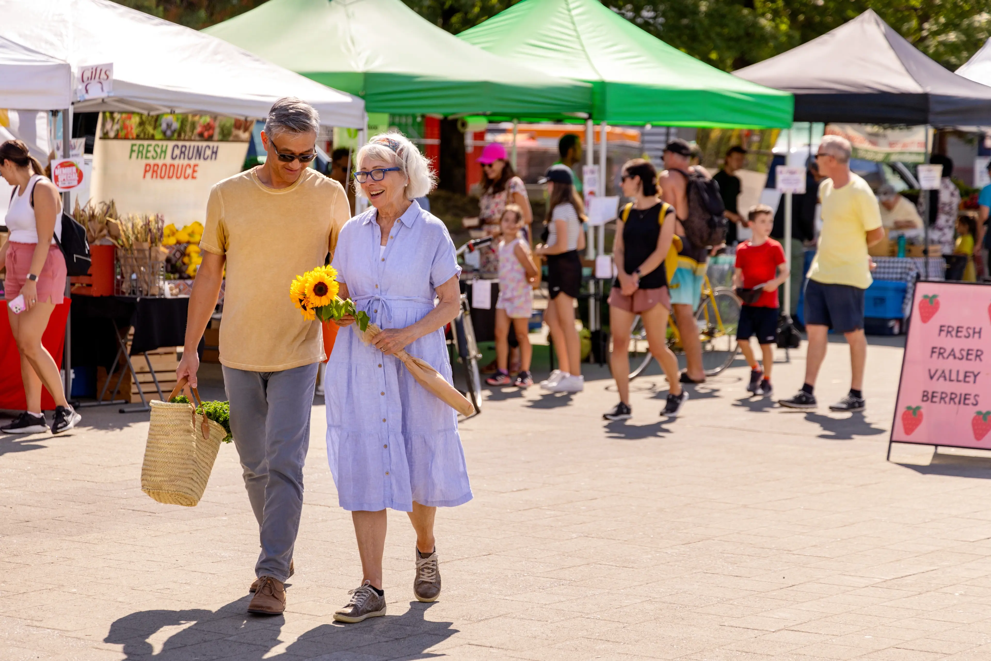 Two people shopping at a farmers' market, one holding sunflowers.