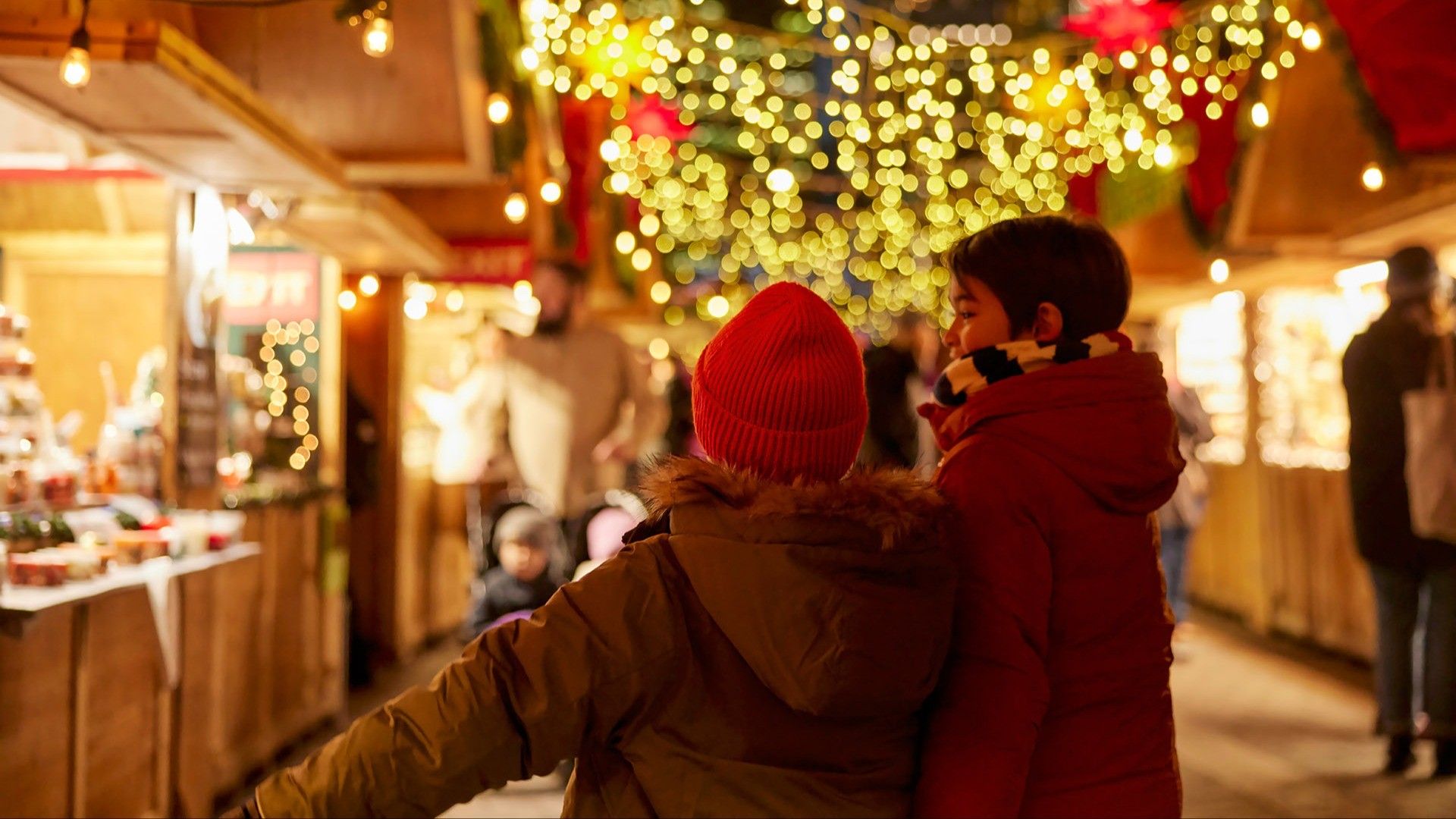 Two children in winter coats and hats walking through a festive market with string lights and wooden stalls.