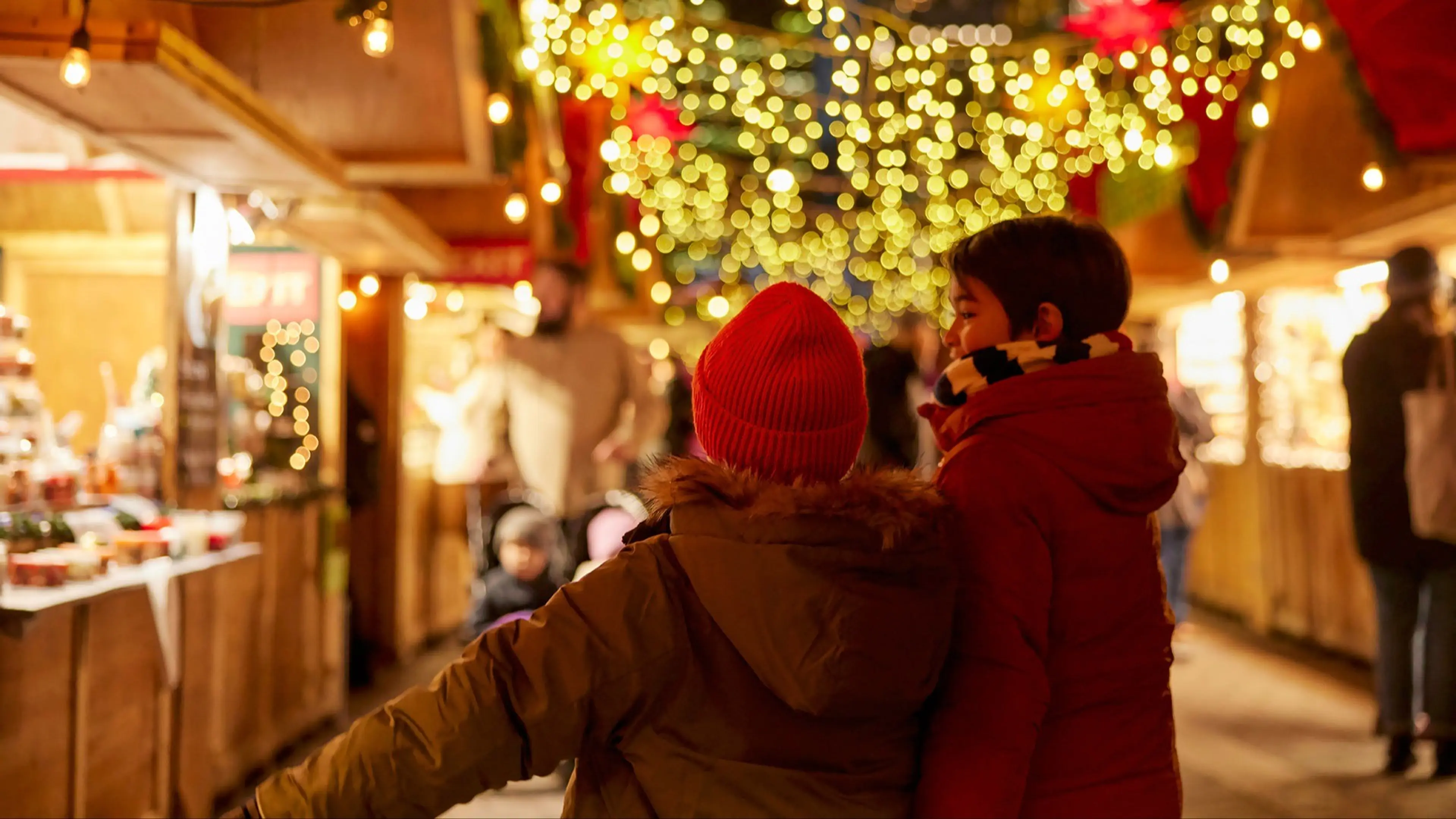 Two children in winter coats and hats walking through a festive market with string lights and wooden stalls.