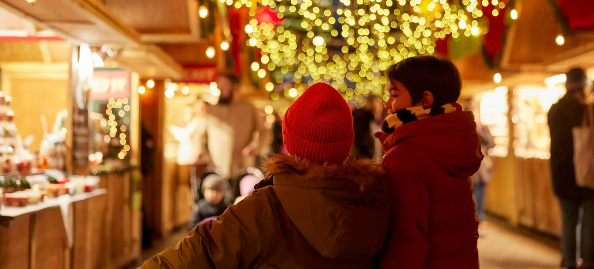 Two children in winter coats and hats walking through a festive market with string lights and wooden stalls.