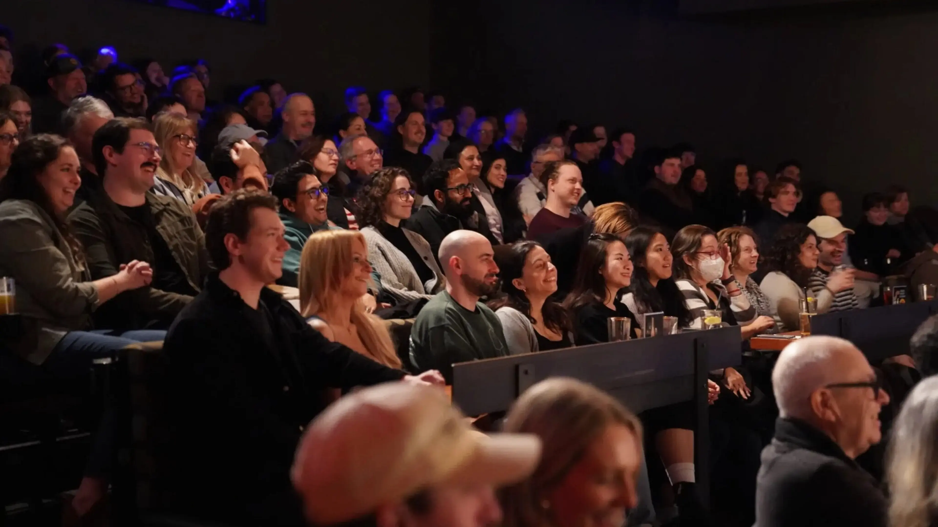 Audience seated in a dimly lit venue, watching an event with smiles and attentive expressions.