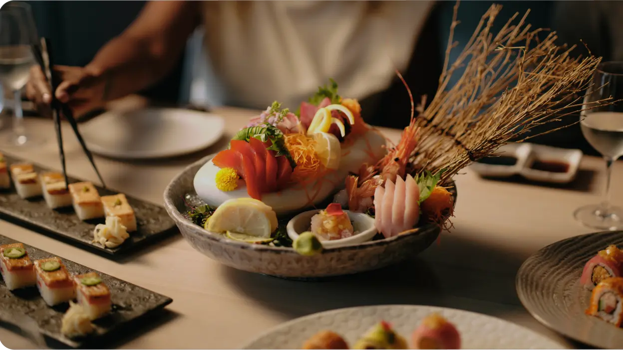 A table set with assorted sushi rolls and a large platter of sashimi, garnished with vegetables and lemon slices—a perfect addition to any Vancouver itinerary. A person is using chopsticks, with glasses of water visible in the background.