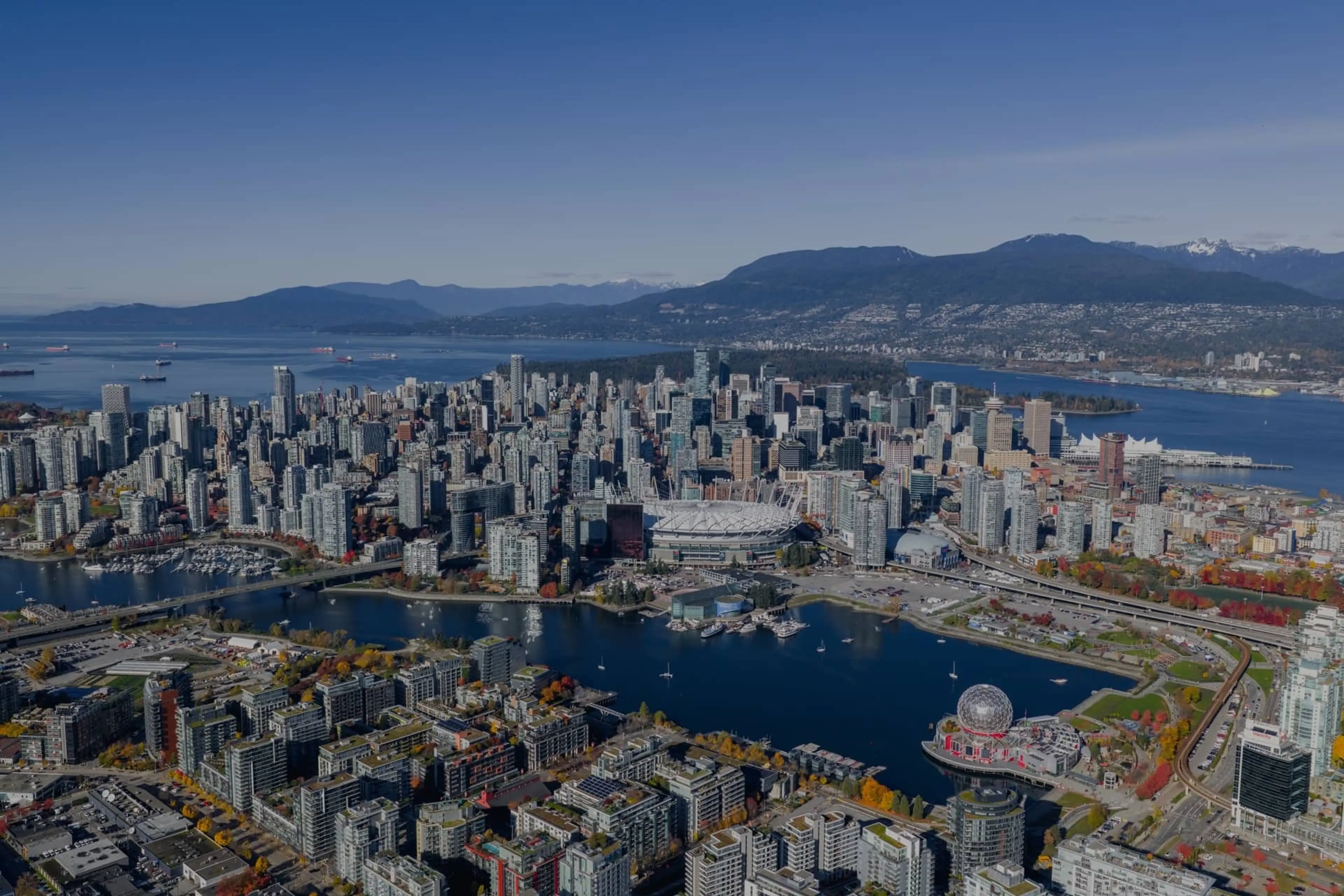 Aerial view of a city with numerous high-rise buildings, a large stadium, water bodies, and mountains in the background under a clear blue sky.