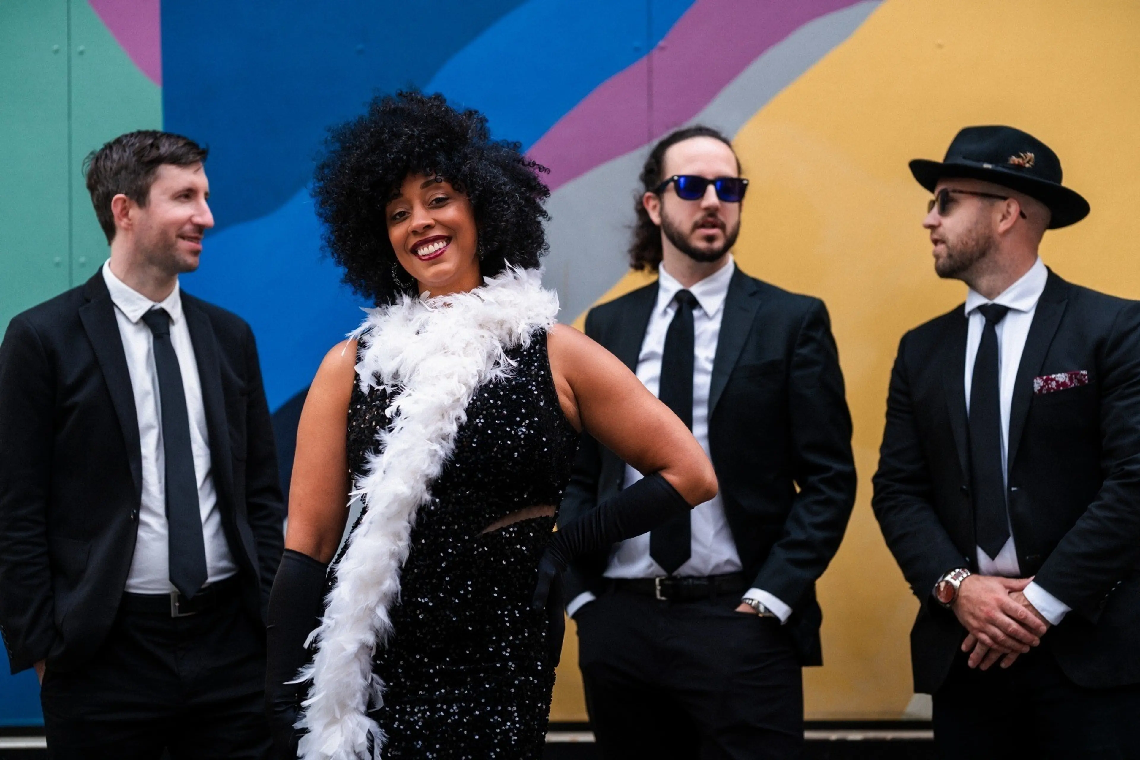 A person in a black sequined dress and white feather boa smiling in front of three people in black suits and ties against a colorful wall.
