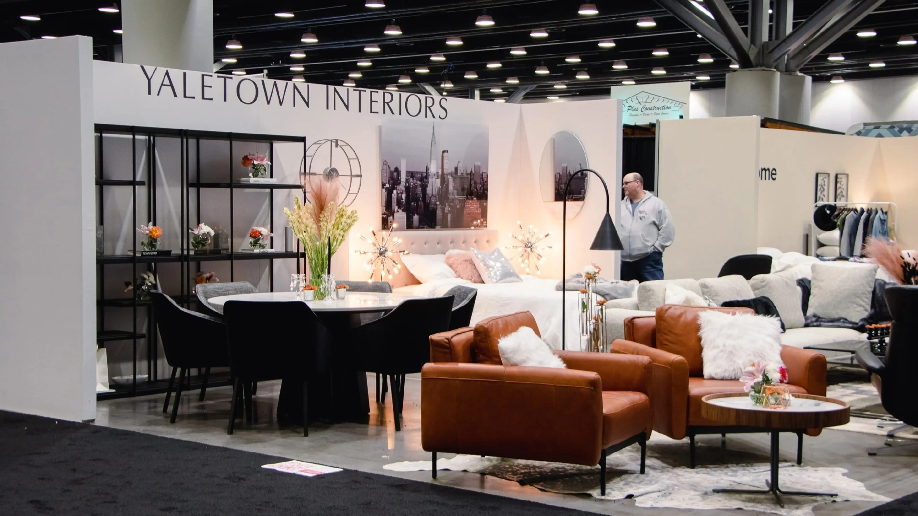 Furniture showroom with brown leather chairs, a round table with black chairs, a bed, and a person standing near a white couch.