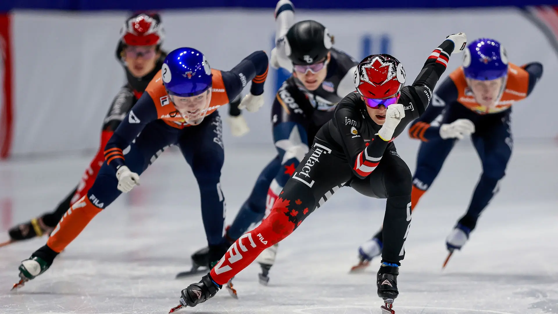 Five speed skaters racing on ice, wearing helmets and racing suits, with one skater in black and red leading.