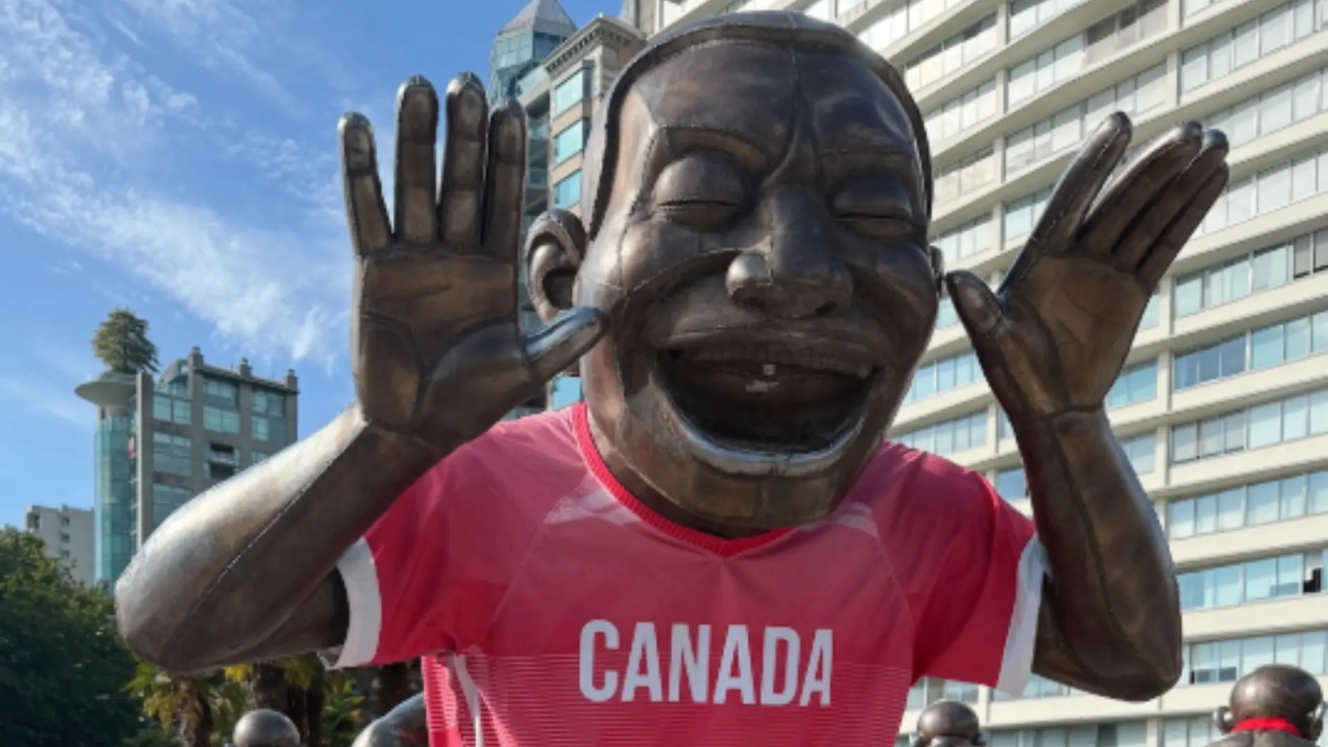 Large bronze statue of a laughing person with hands raised near the face, wearing a red Canada shirt, in an urban setting.