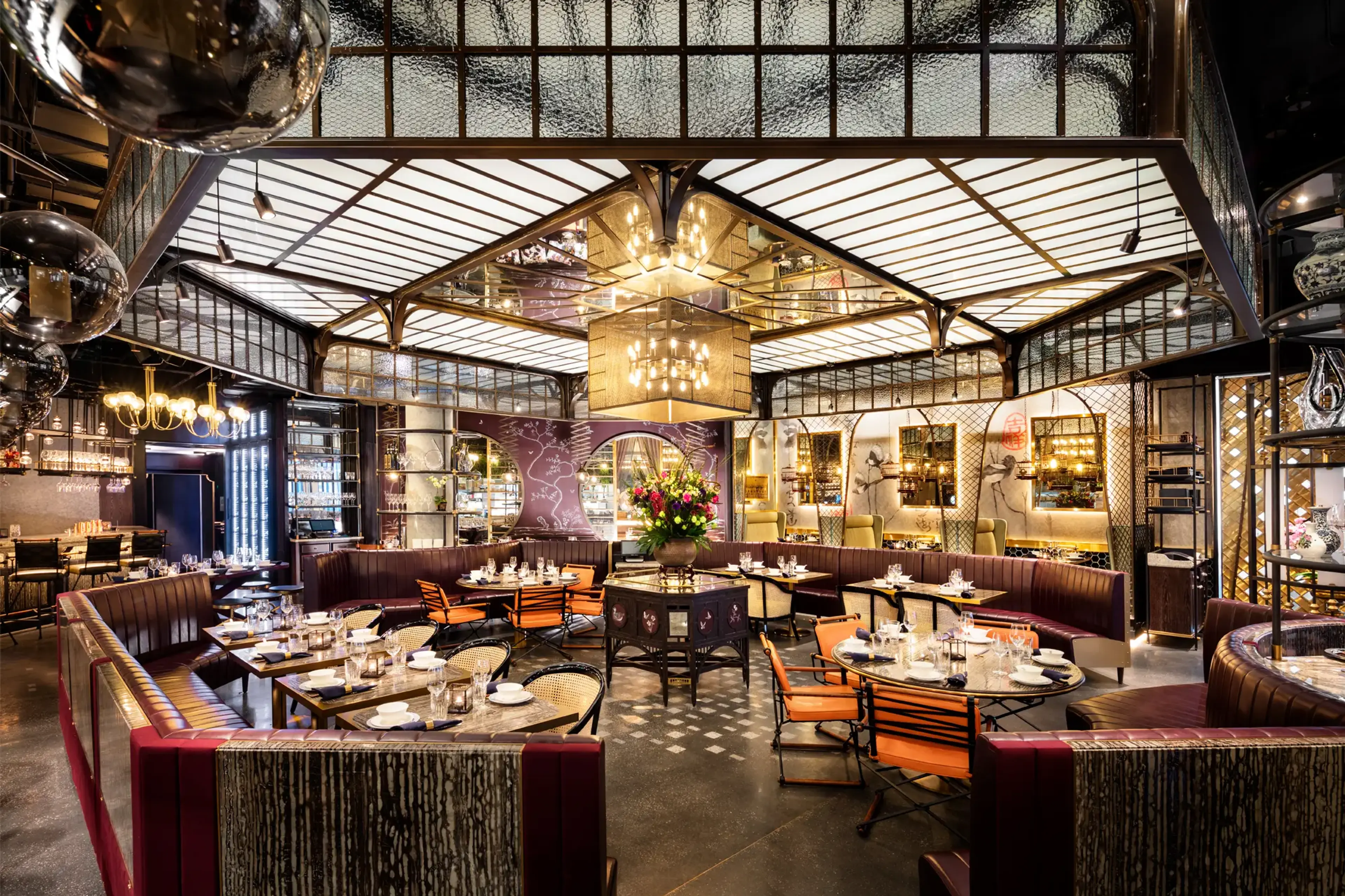 Upscale restaurant interior with curved leather booths, round tables set with glassware, and a central floral arrangement under a large ceiling light.