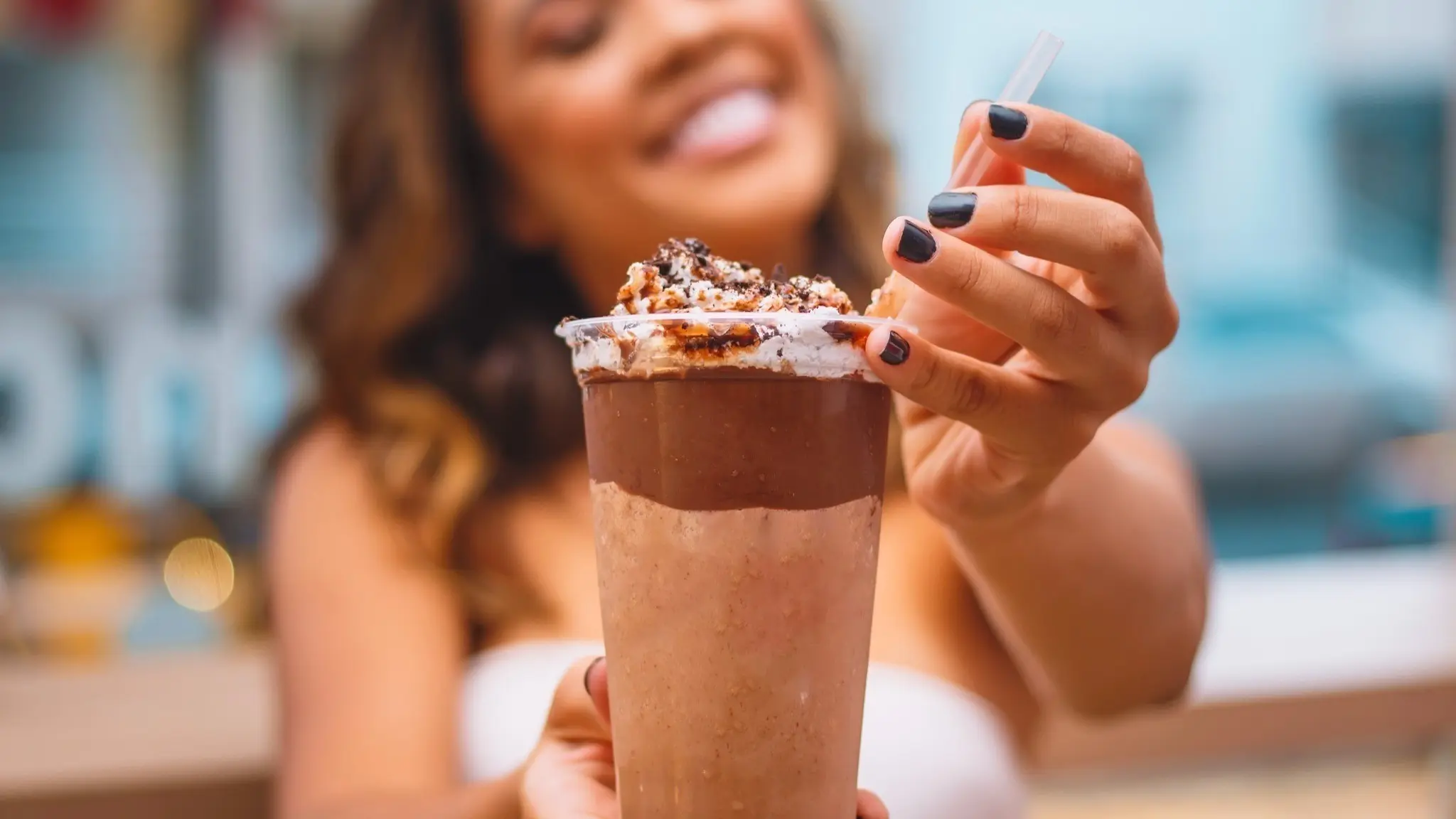 Person holding a chocolate milkshake topped with whipped cream and chocolate pieces, smiling in the background.