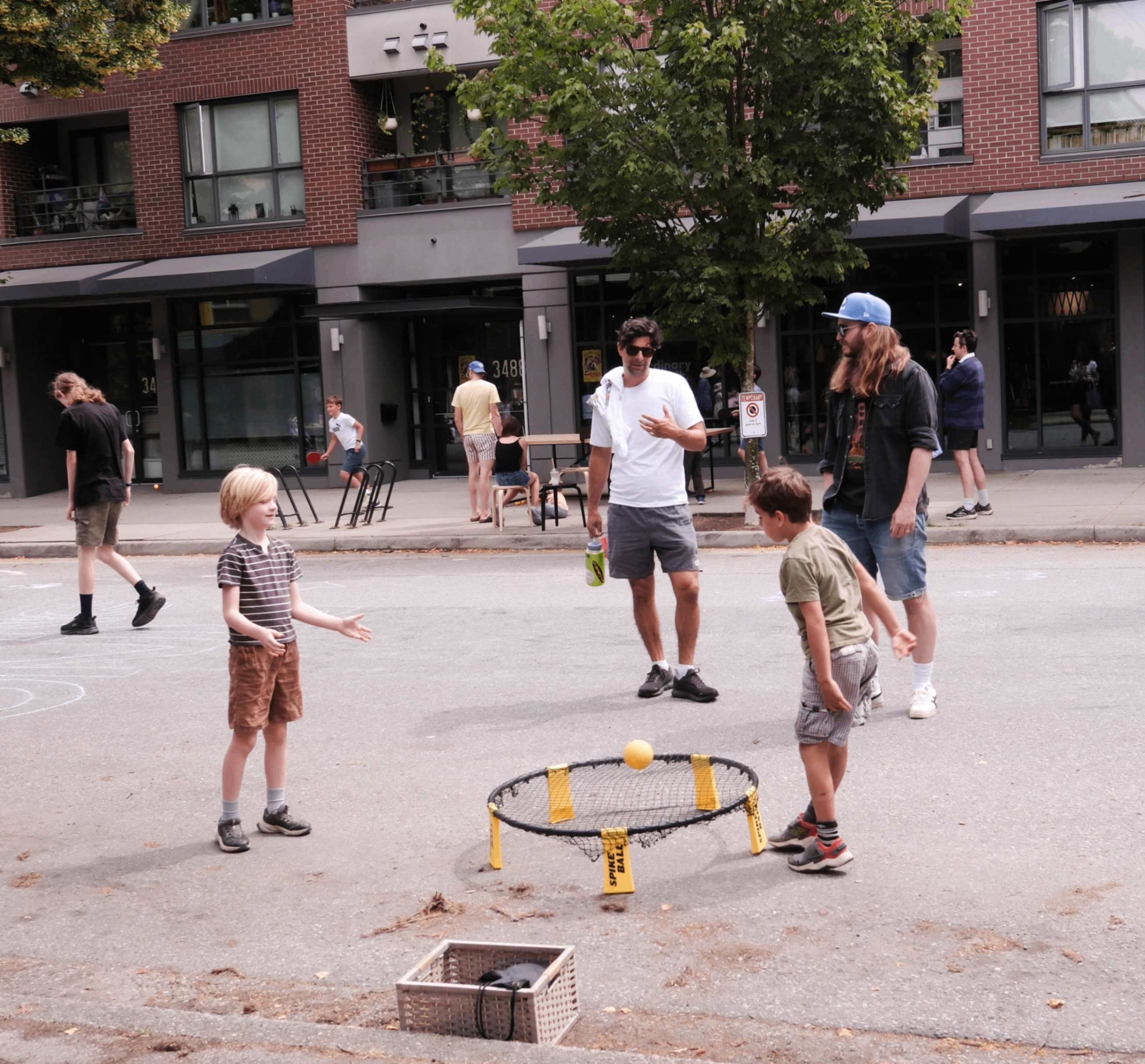 Two children playing spikeball on a street with two adults watching nearby and other people in the background.
