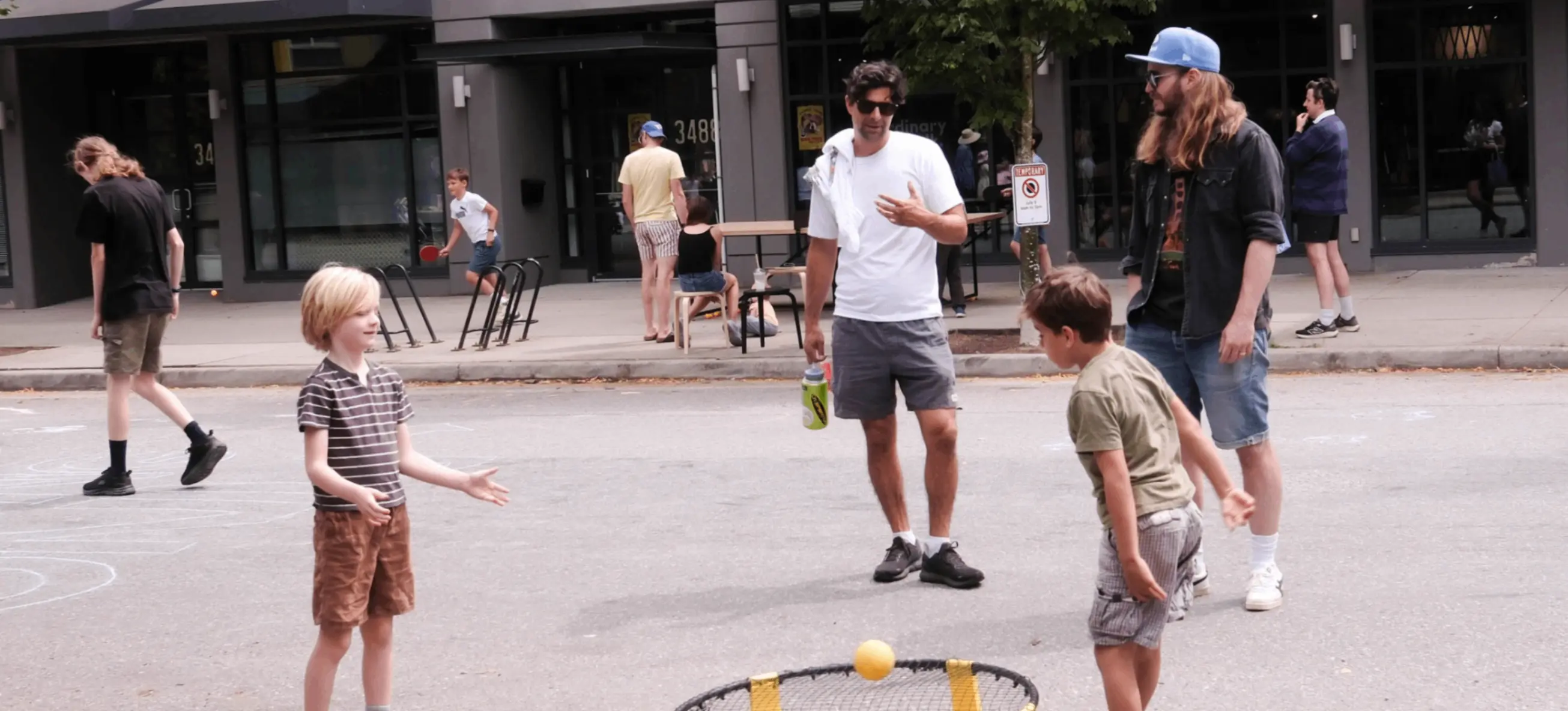 Two children playing spikeball on a street with two adults watching nearby and other people in the background.
