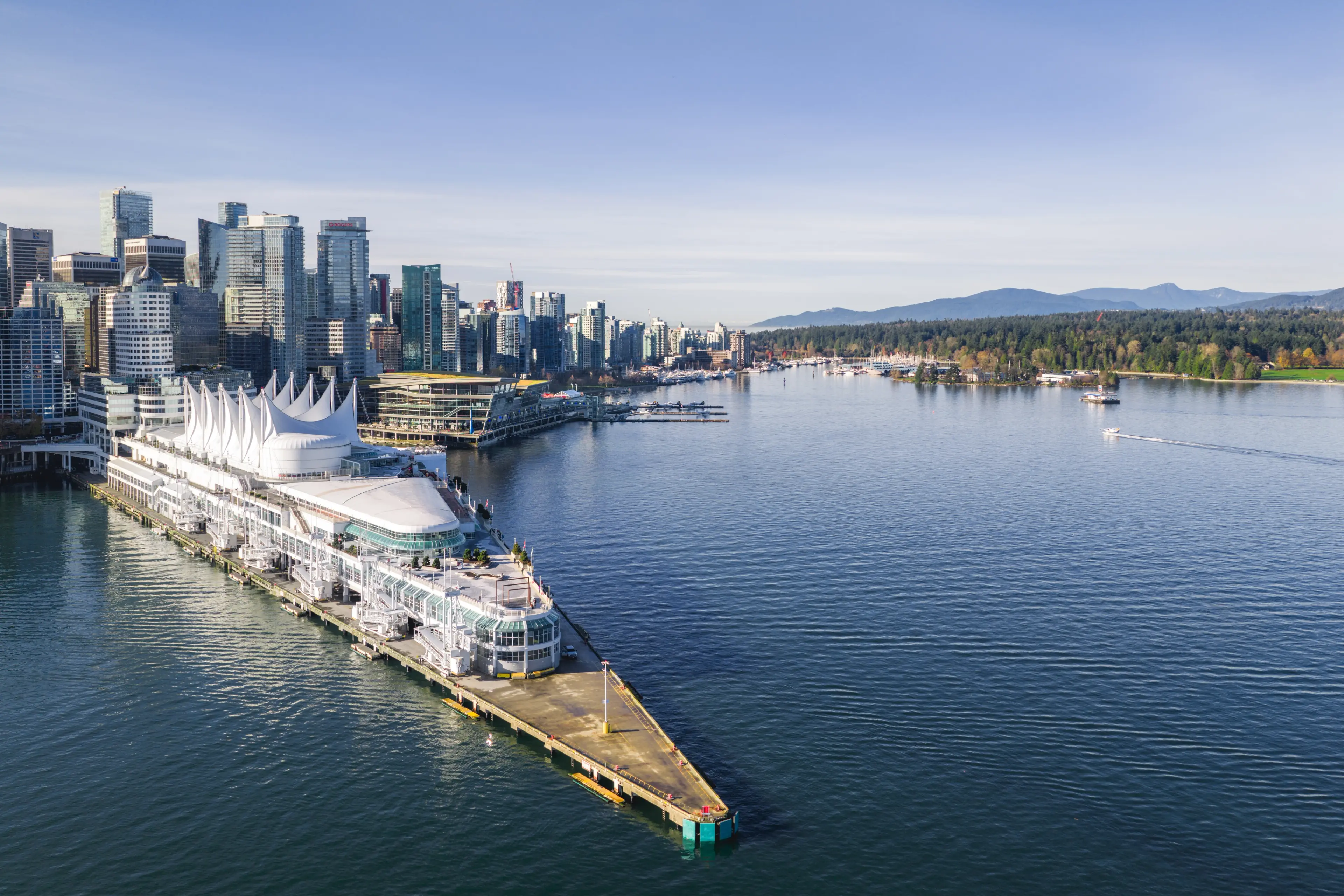A waterfront city skyline with modern high-rise buildings and a large white building with sail-like structures on a pier extending into calm water.