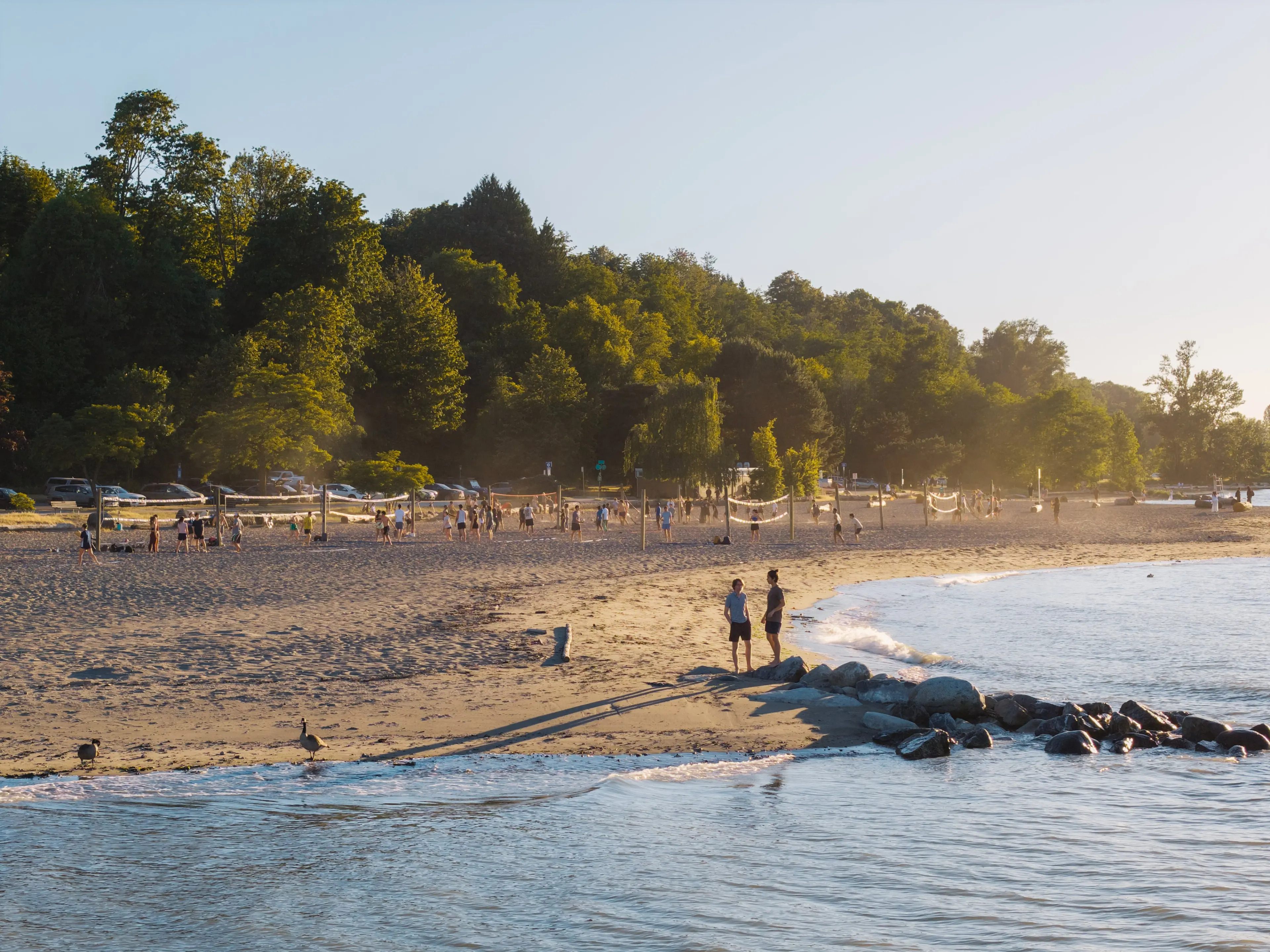 Two people standing on a sandy beach near rocks with volleyball nets and many people playing in the background under trees.