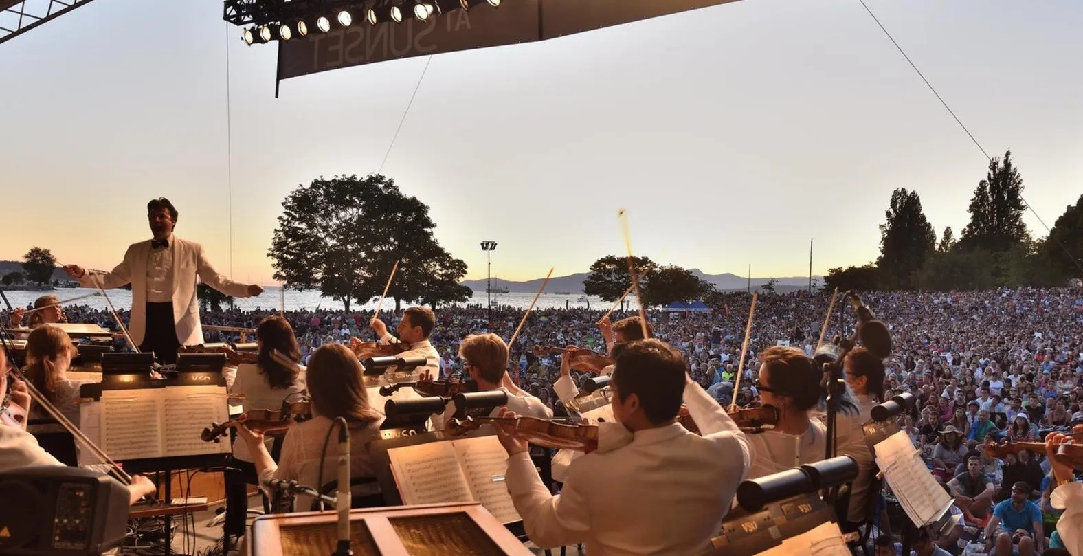 Orchestra performing outdoors at sunset with conductor leading and a large audience in the background.