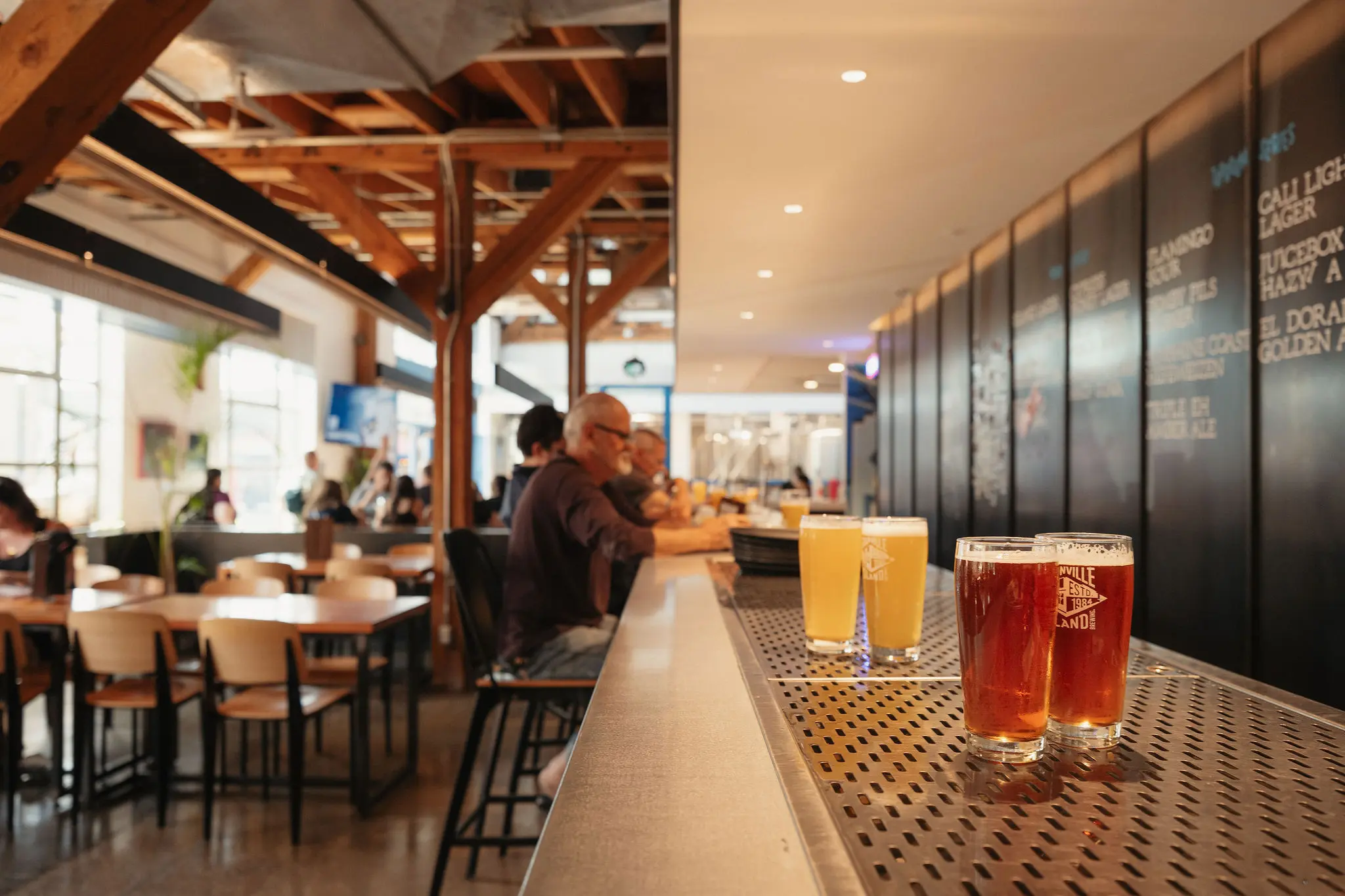 Four glasses of beer on a bar counter with people sitting and talking in the background of a brewery or pub.