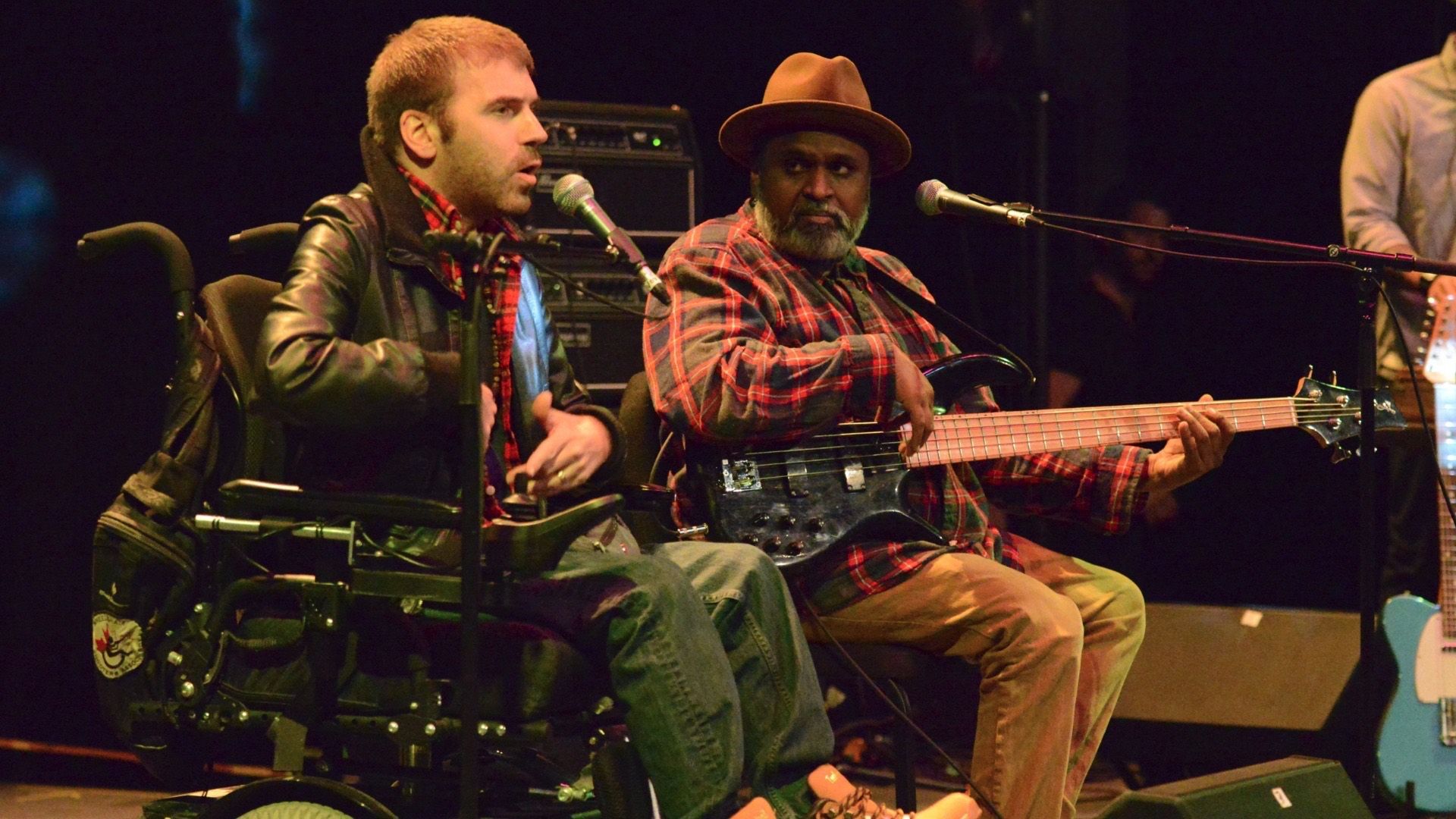 Two people on stage, one in a wheelchair speaking into a microphone, the other playing an electric bass guitar.