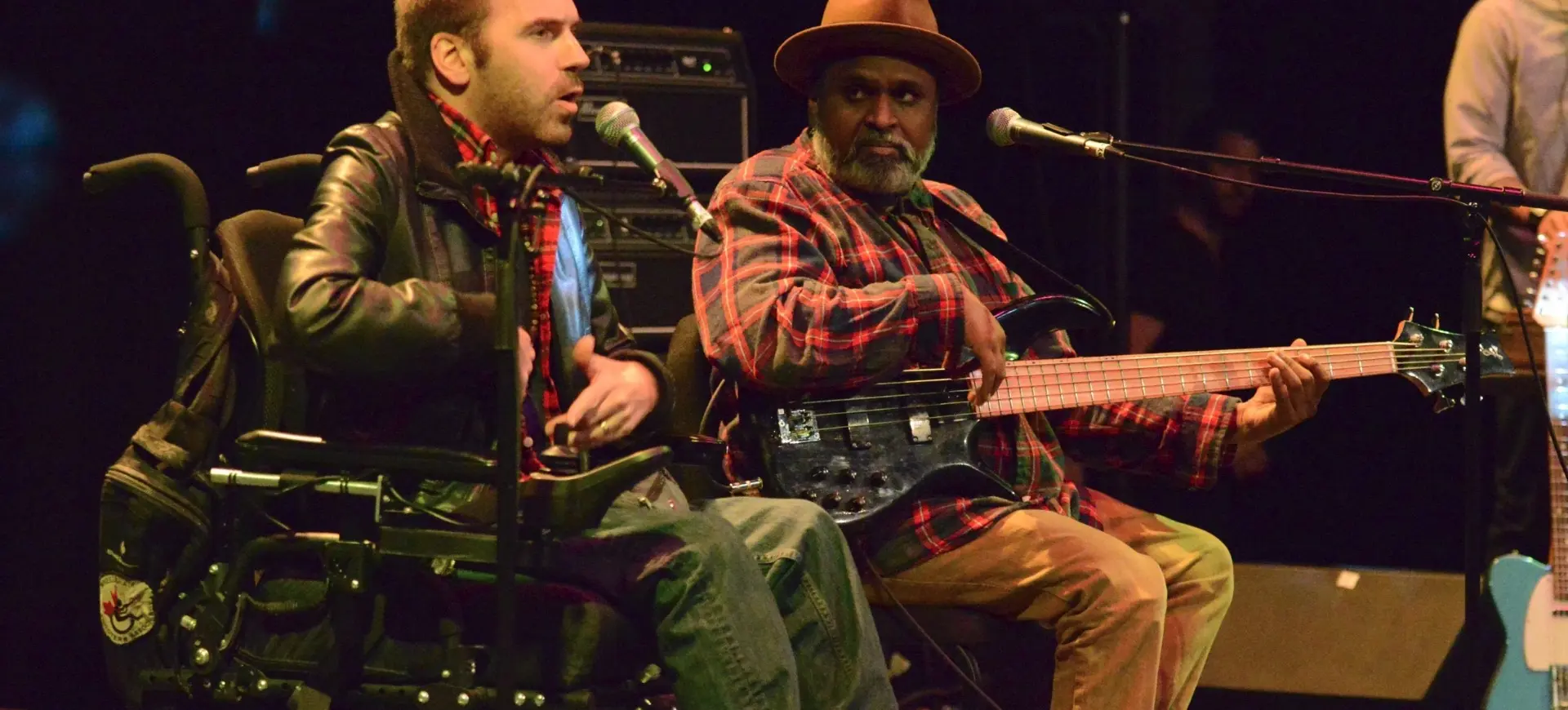 Two people on stage, one in a wheelchair speaking into a microphone, the other playing an electric bass guitar.
