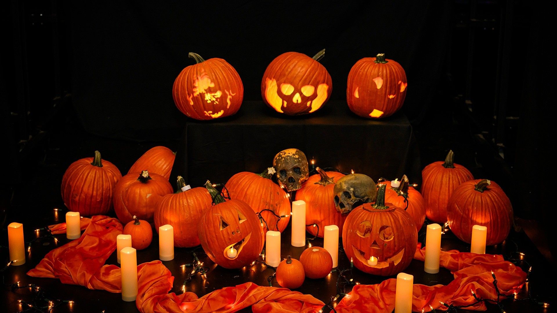 Group of carved and uncarved pumpkins with lit candles, skull decorations, and orange fabric on a dark background.