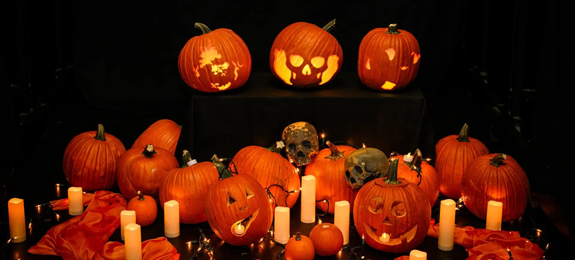 Group of carved and uncarved pumpkins with lit candles, skull decorations, and orange fabric on a dark background.