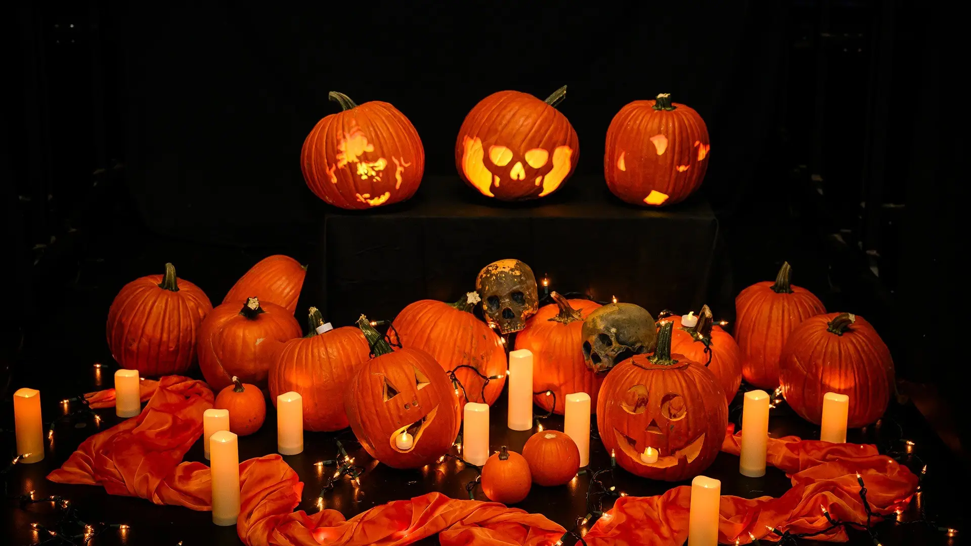 Group of carved and uncarved pumpkins with lit candles, skull decorations, and orange fabric on a dark background.