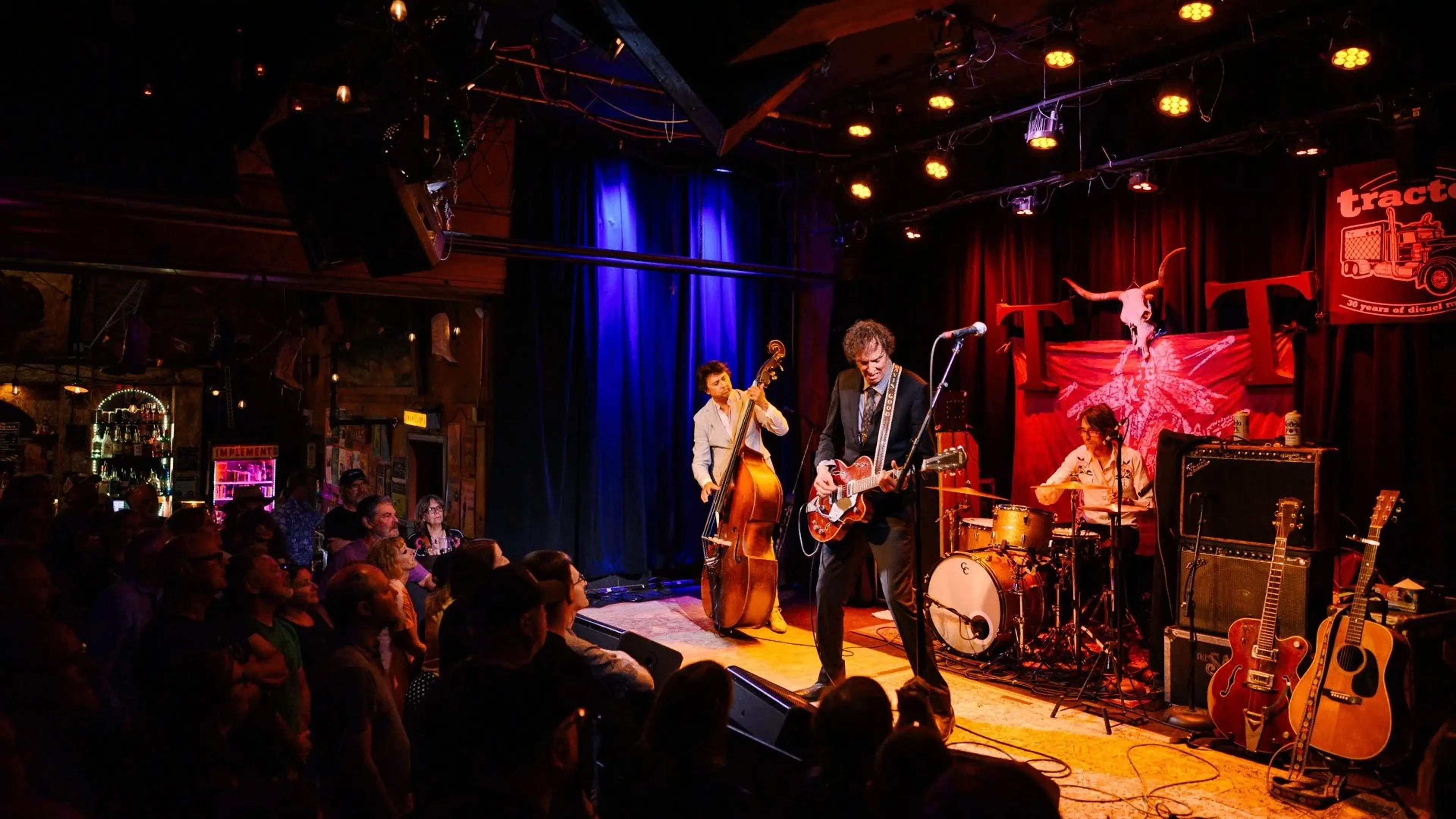 A band performs on stage with a guitarist, upright bassist, and drummer in front of an audience in a dimly lit venue.